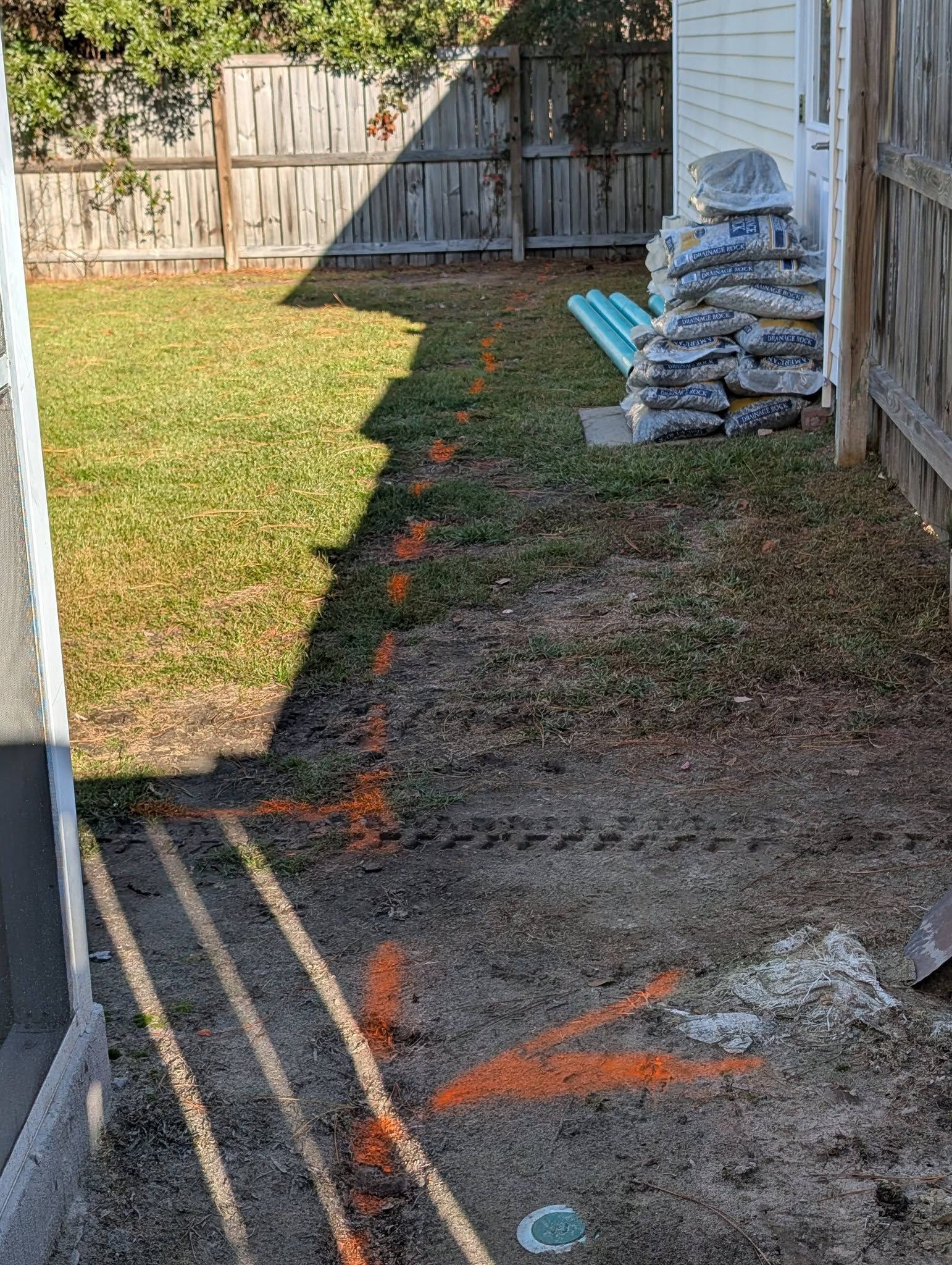 Narrow backyard with grass, dirt strip, stacked materials, and orange spray markings along the ground and fence.