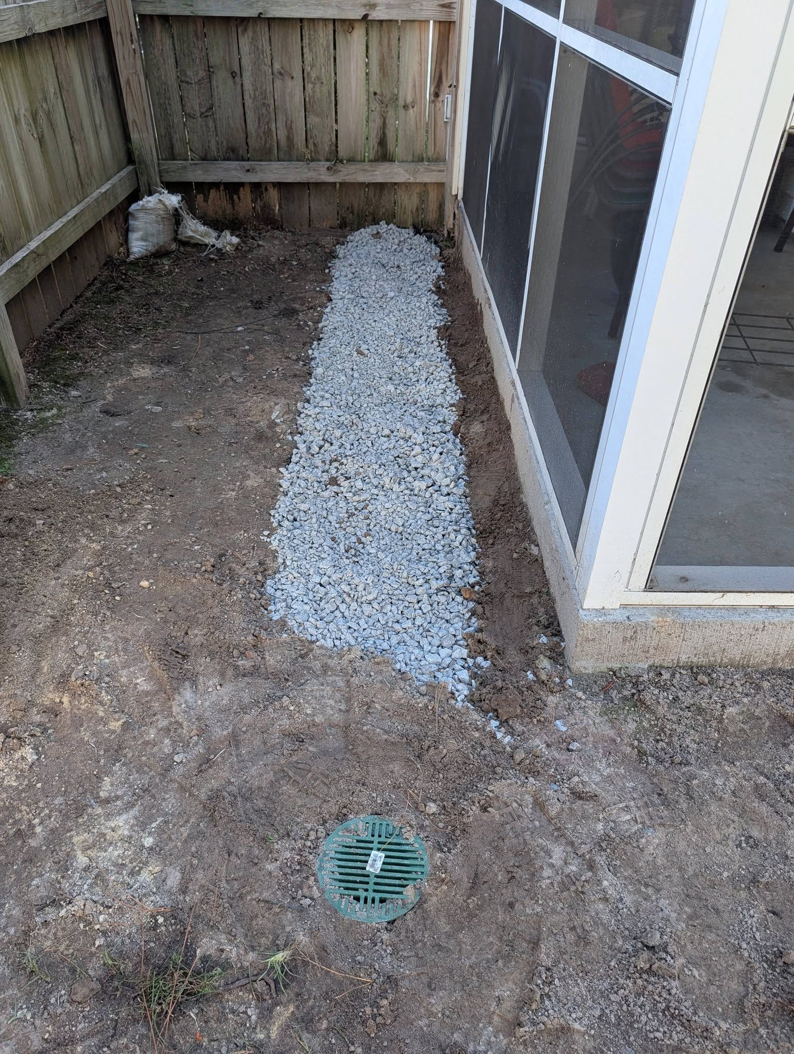 Narrow outdoor side yard with gravel, a drain, and a screened door beside a wooden fence.