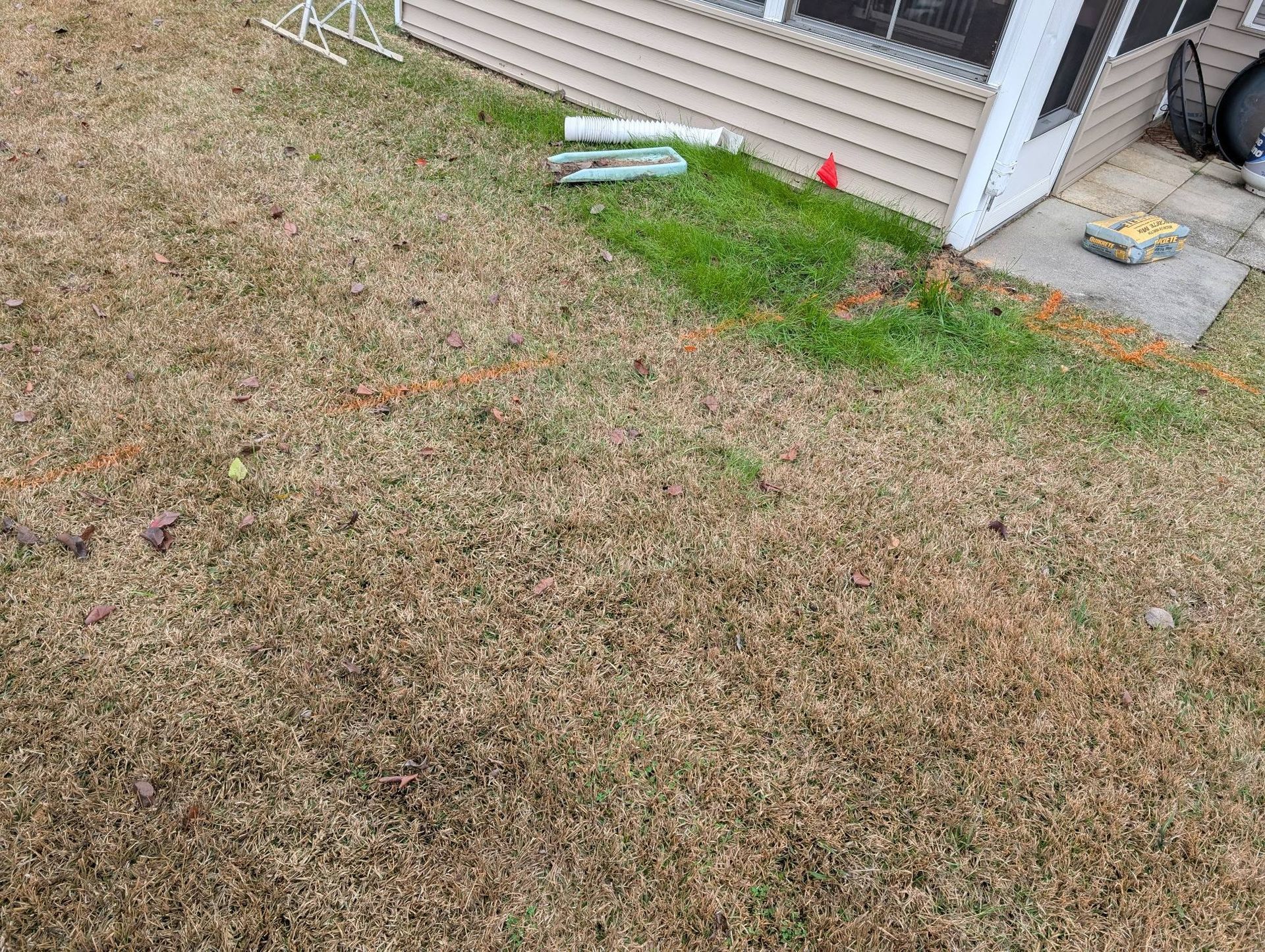 Patchy brown lawn beside a house porch, with a strip of green grass and a few small items near the steps