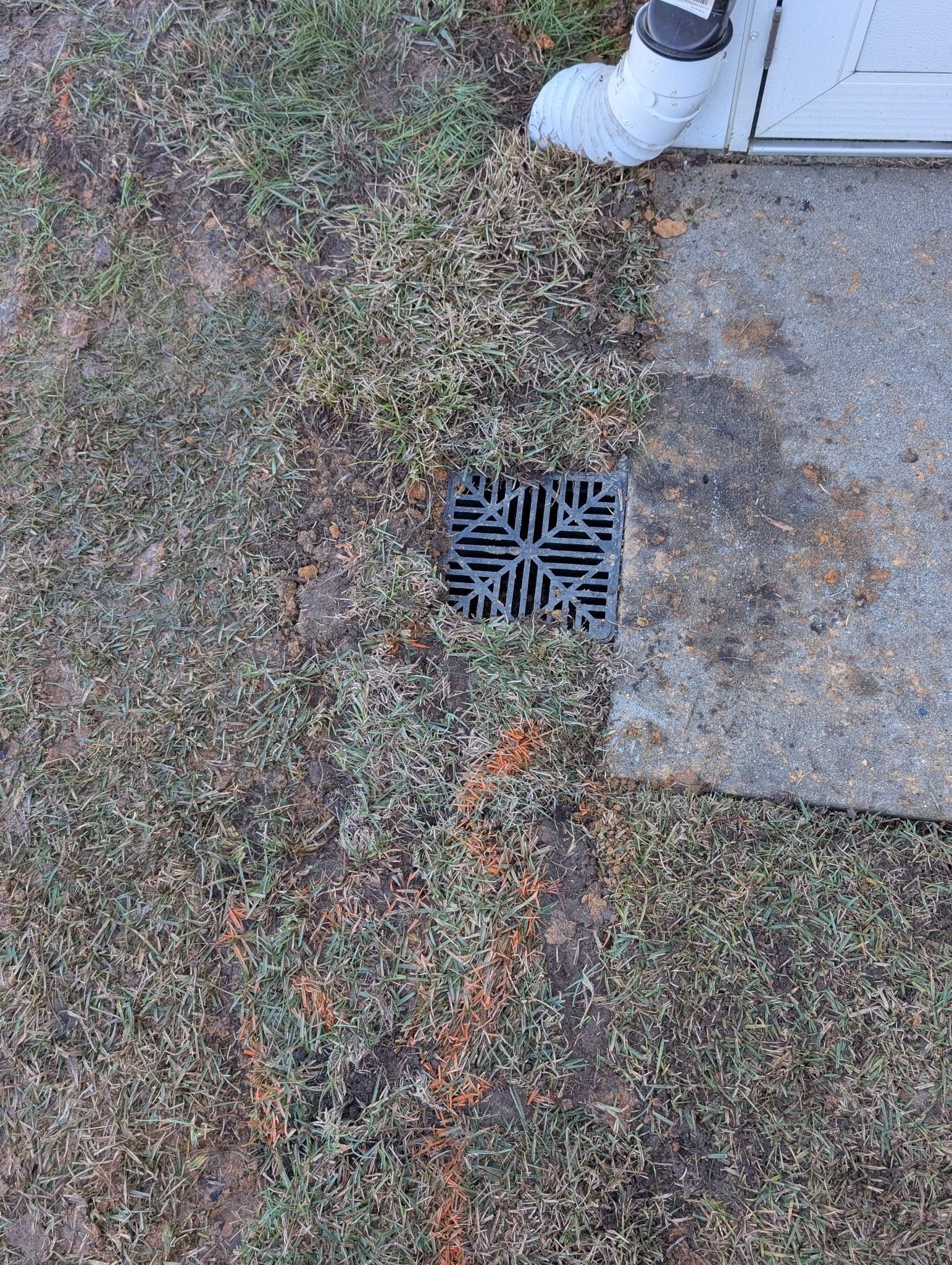 Drain grate beside a concrete path, with a person’s white sneaker at the top edge.