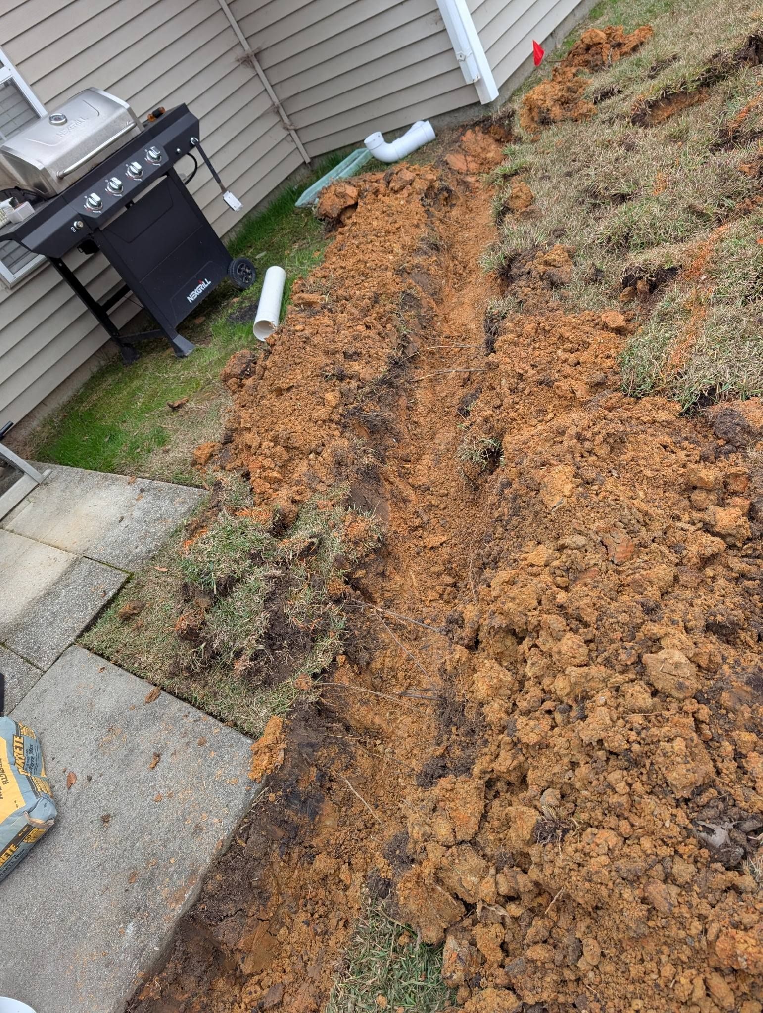 Backyard trench with exposed orange-brown soil beside a patio and house siding.
