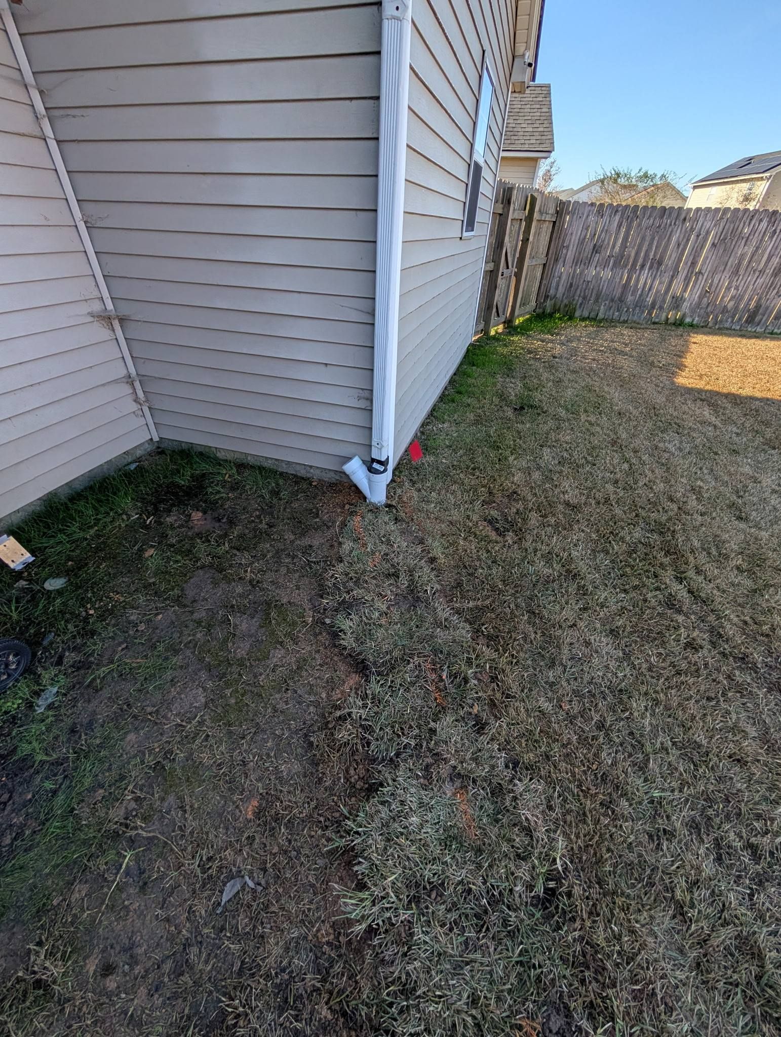 Side of a house with siding, a downspout, and a narrow dirt strip beside a fenced yard.