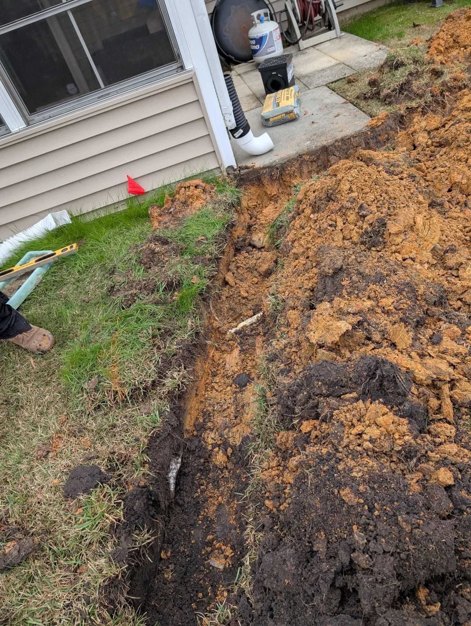 Large muddy trench beside a house, with orange soil and a small red marker flag along the edge