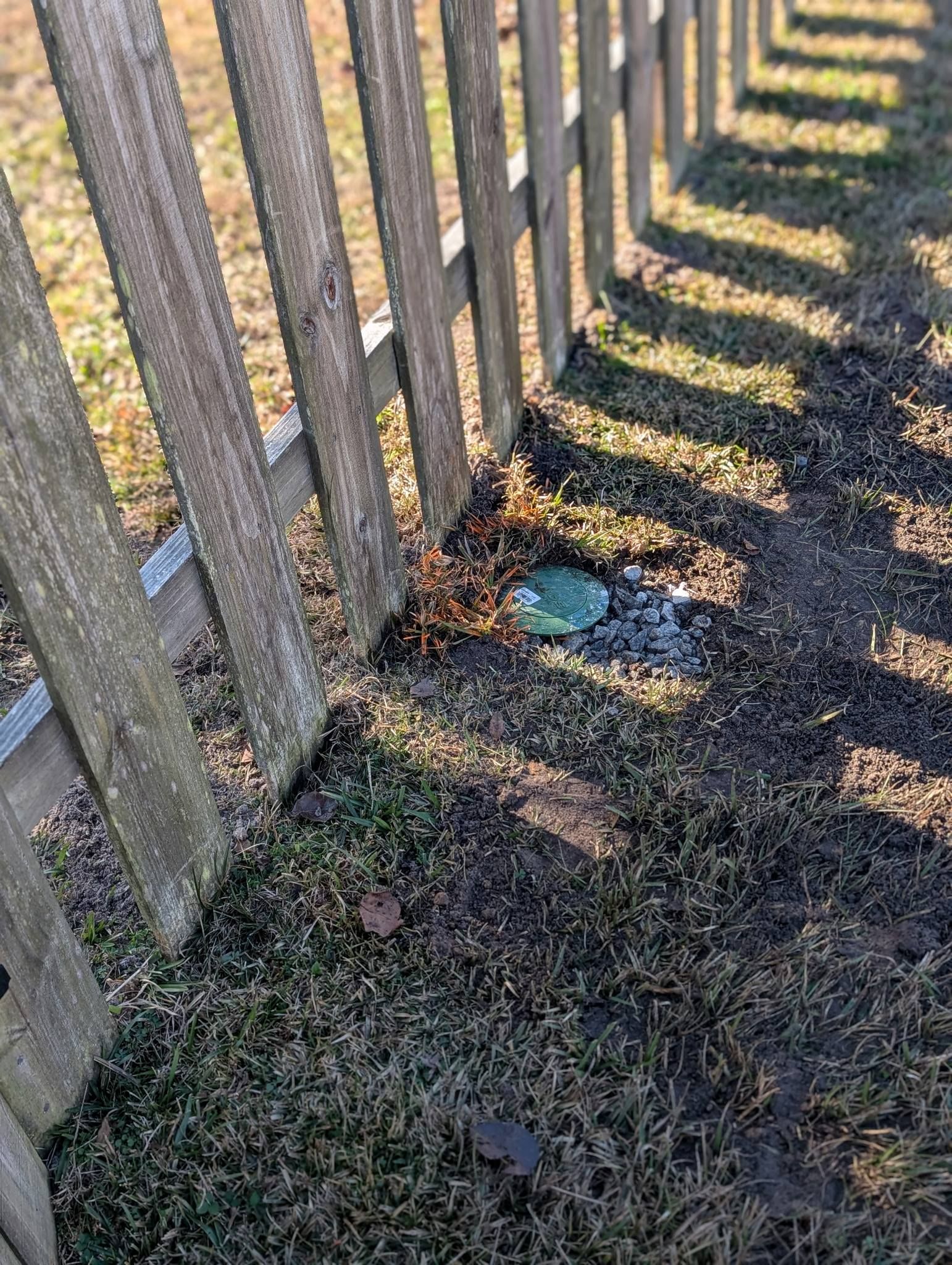 Sunlit wooden fence casting shadows on a gravel path with patchy grass and a small blue object on the ground