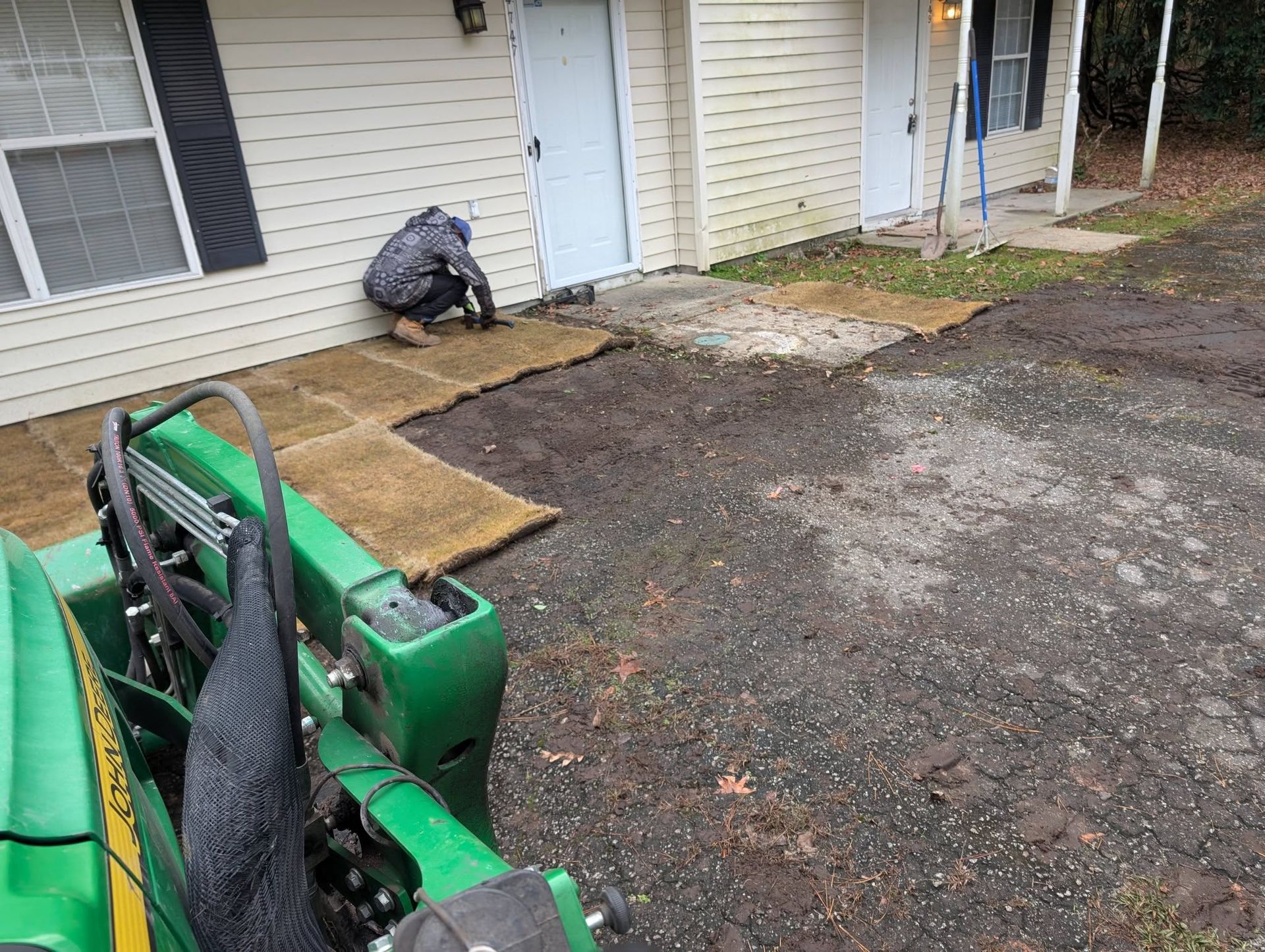 Green tractor loader in a muddy driveway beside a house with plywood and concrete pads near the entrance