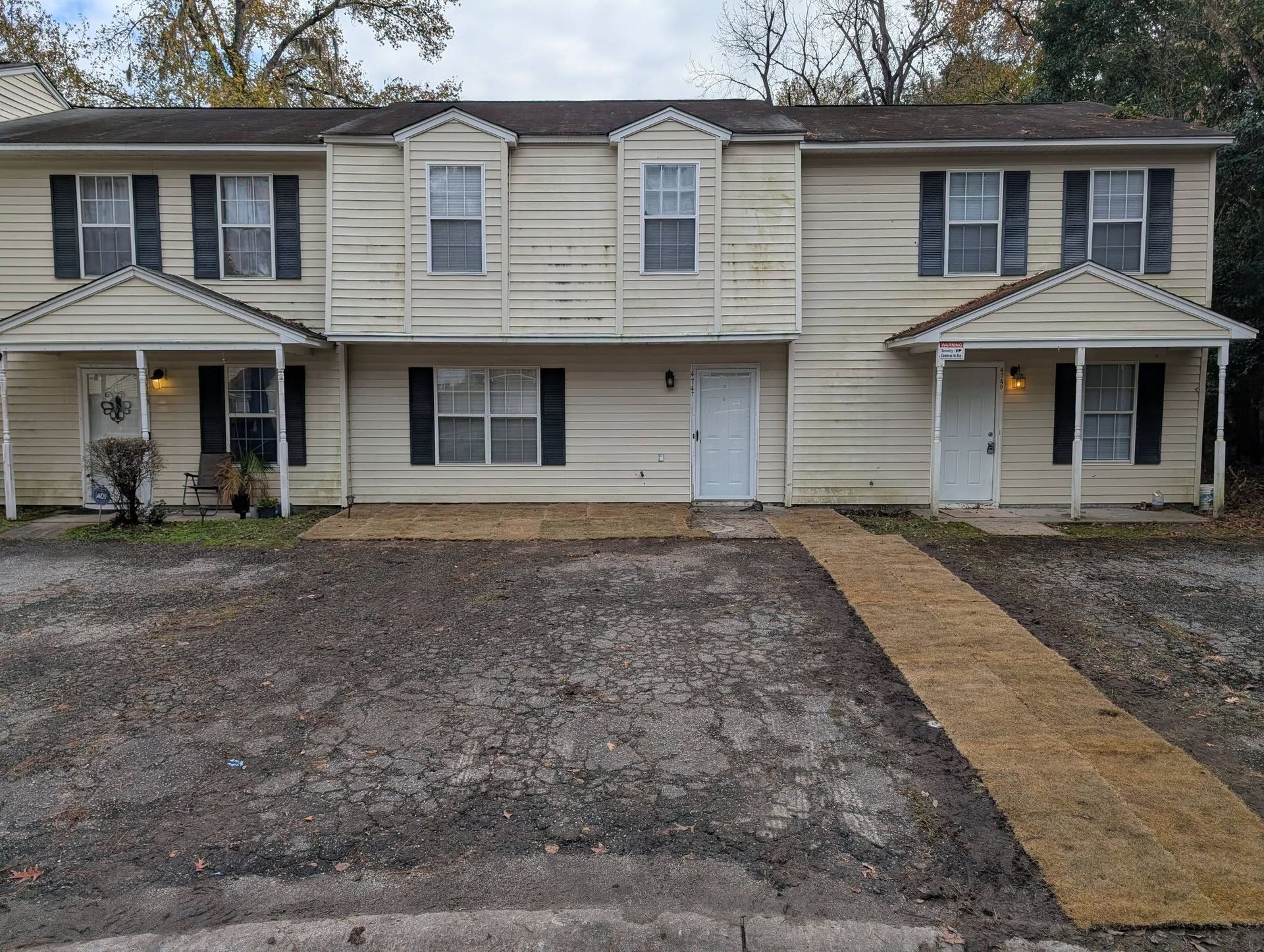 Two-story duplex with light siding, dark shutters, and a gravel front yard with a narrow walkway