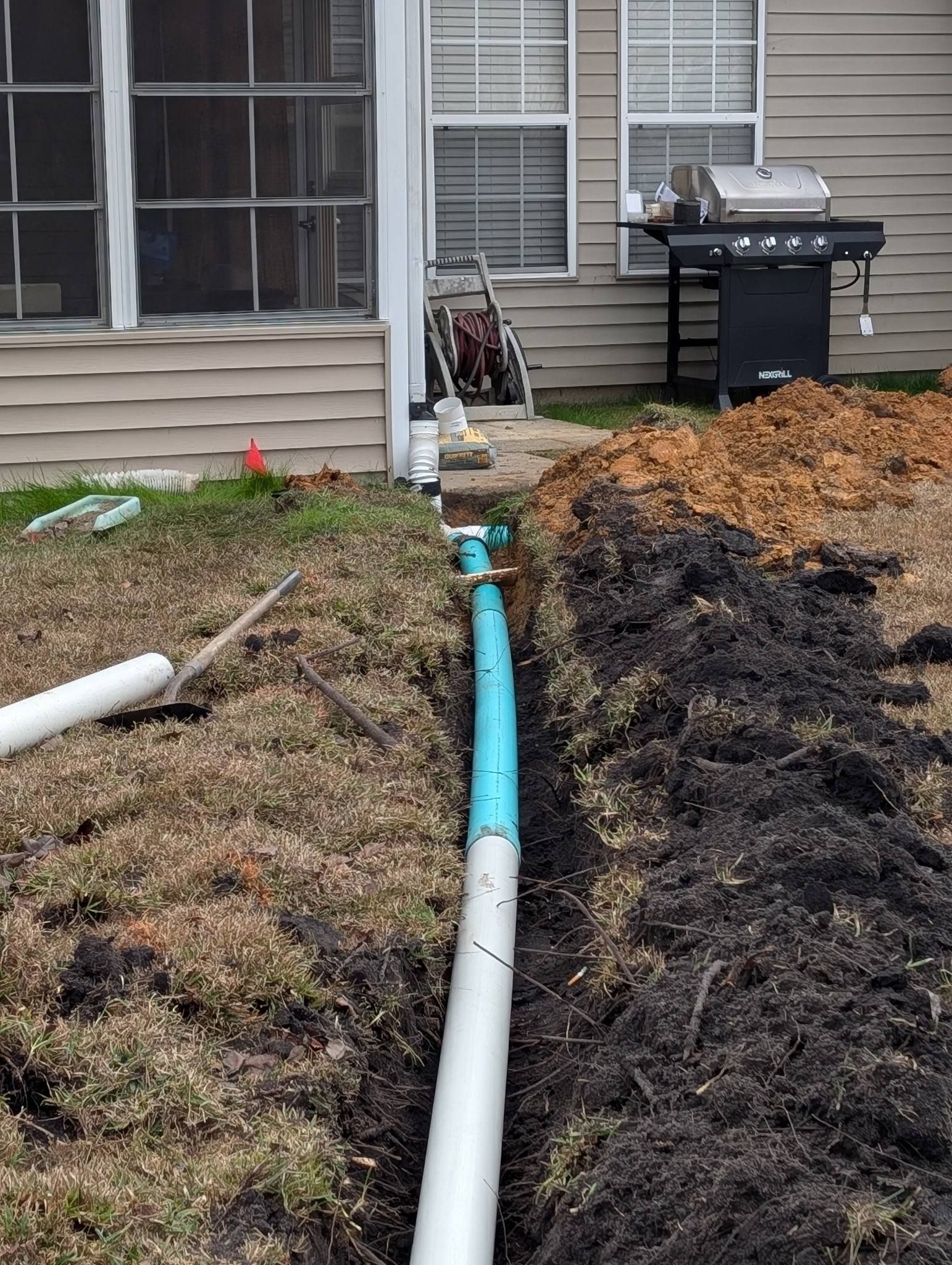 Backyard with a blue pipe trench beside a house and grill, lined with mulch and soil piles