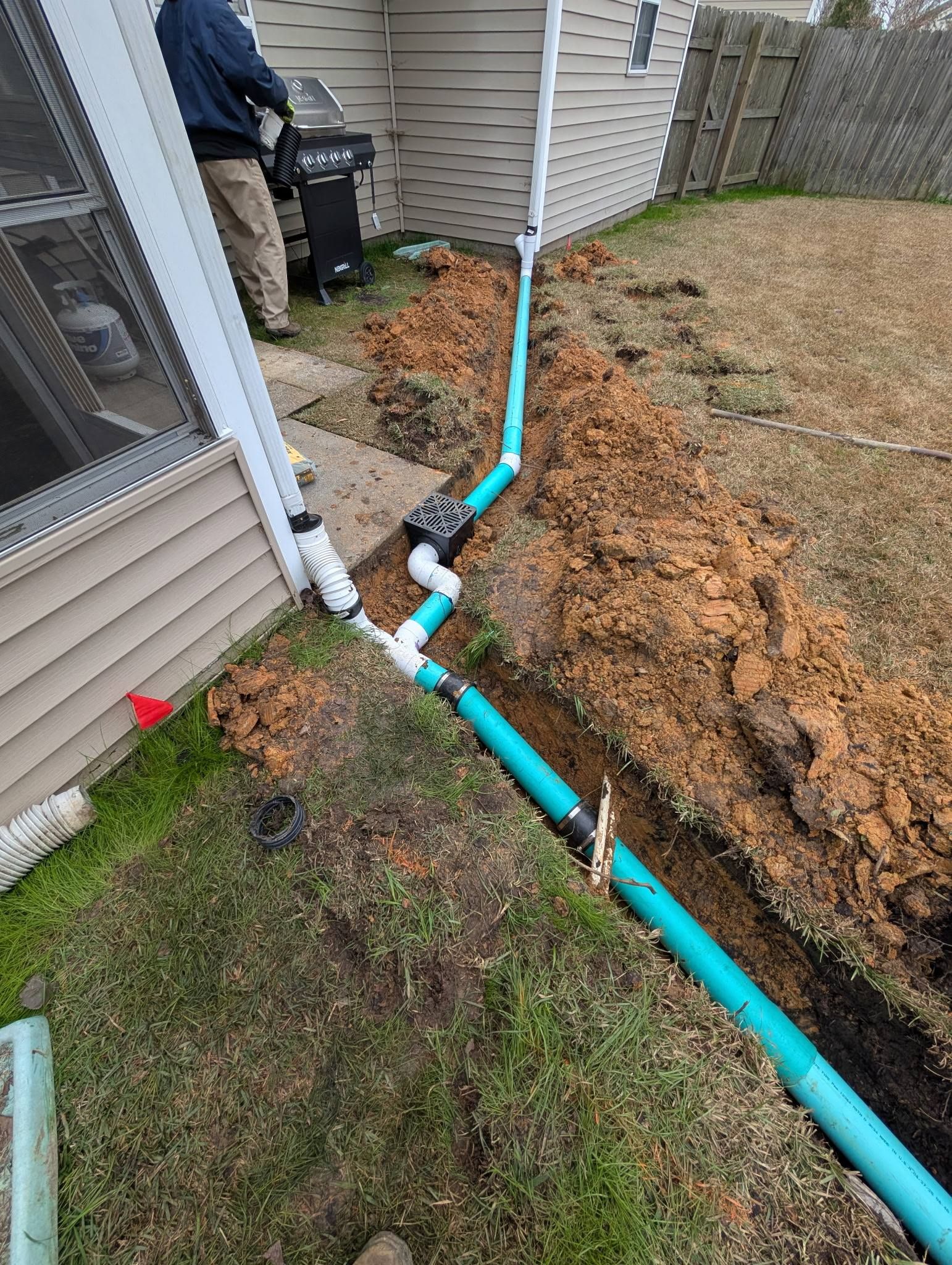 Blue drainage pipe installed beside house in a muddy trench next to a fenced yard.