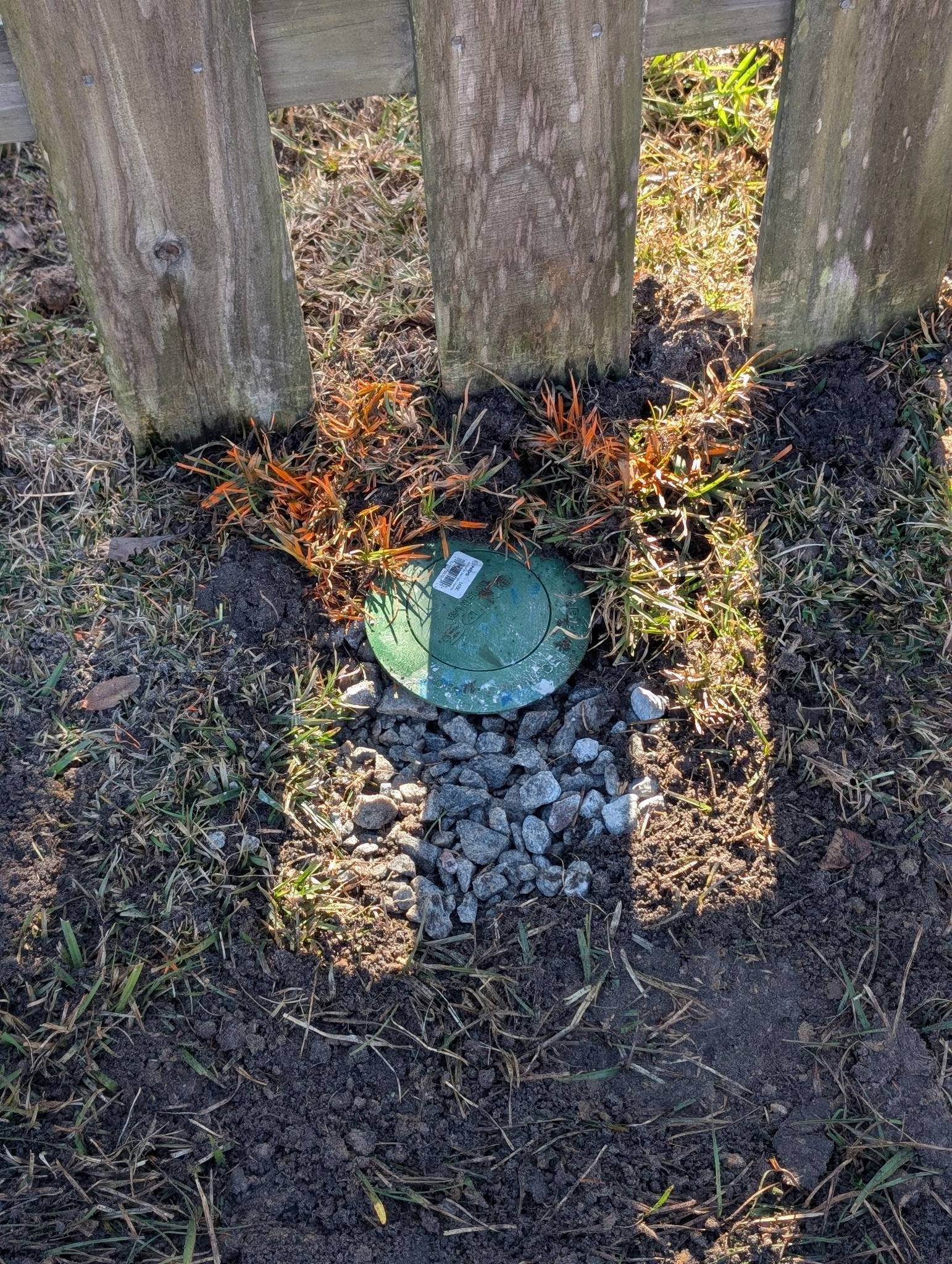 A green utility meter cover set in gravel beside a wooden fence, with fallen leaves around it.