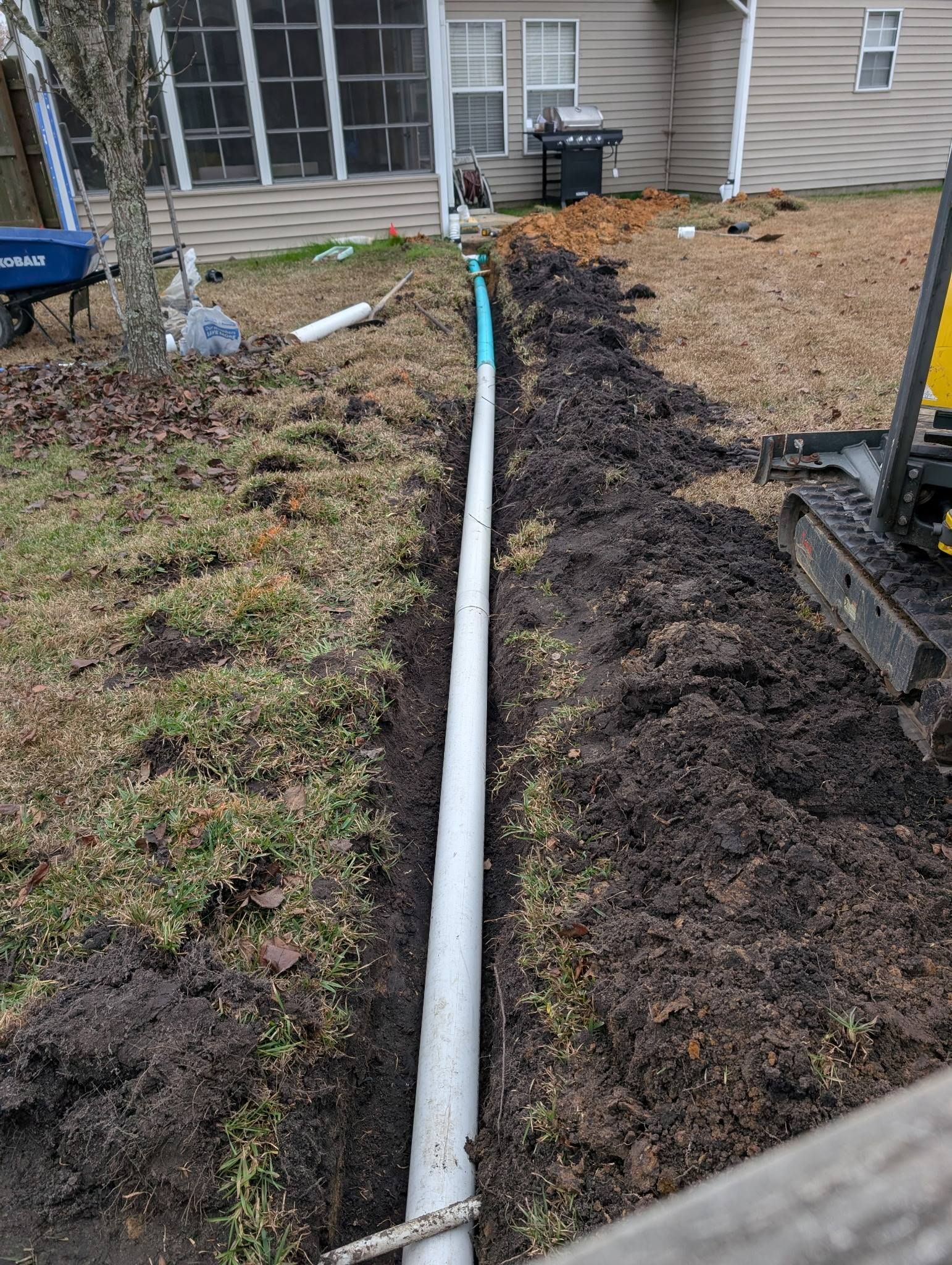 Yard trench with white pipe running to a house, bordered by dirt and patchy grass.