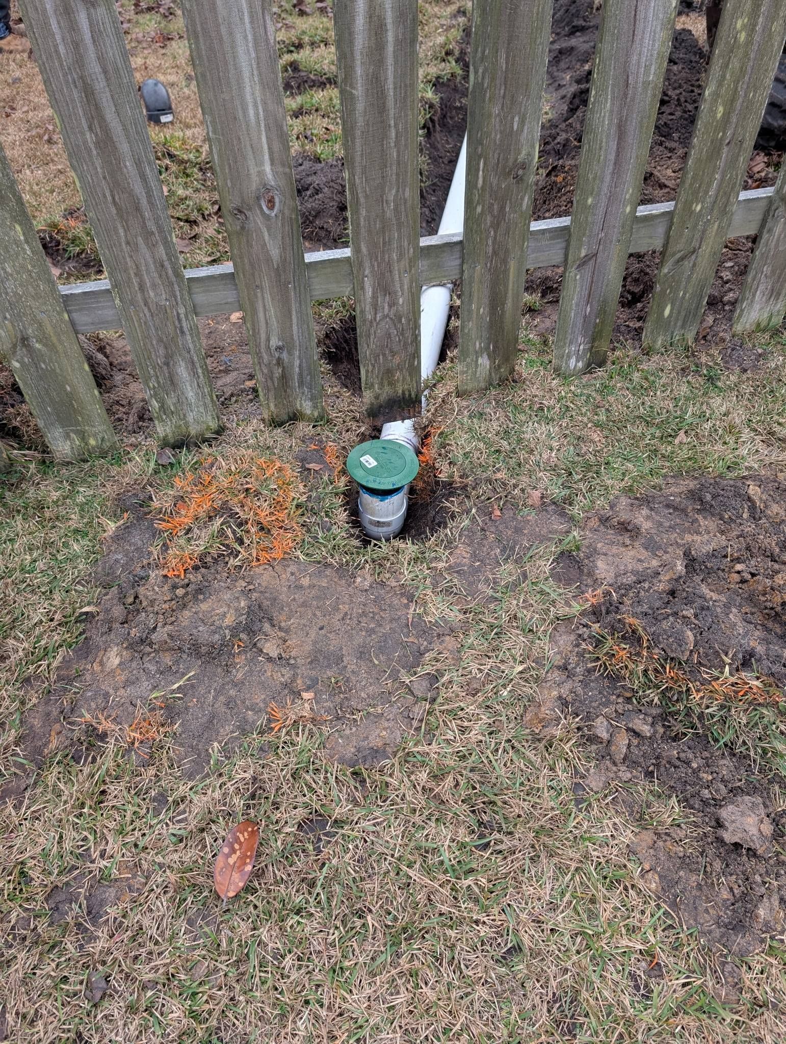 Small green utility box beside a wooden fence, surrounded by muddy ground and grass.