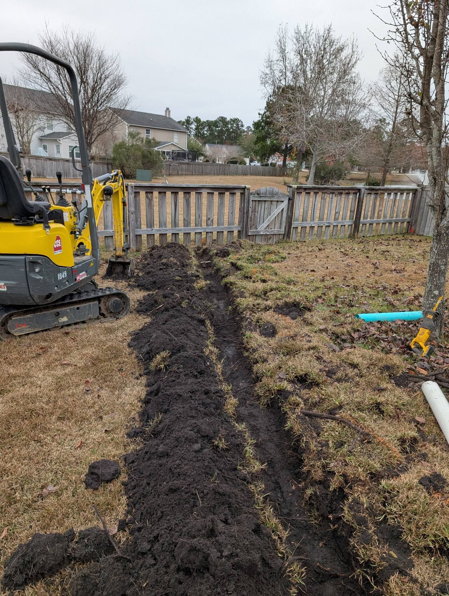 Narrow trench cut through a backyard lawn beside a yellow excavator, with a fence in the background