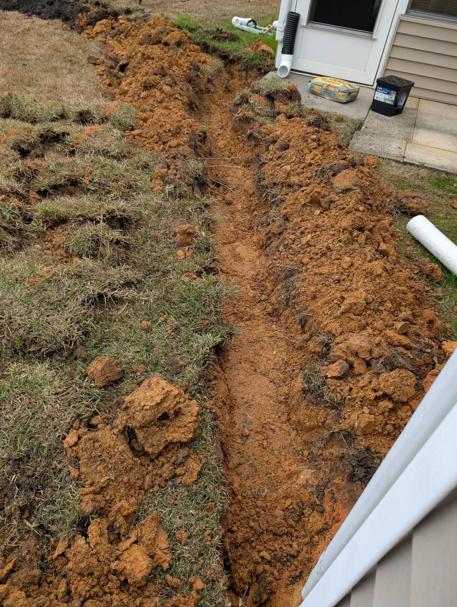 Freshly dug trench in muddy soil beside a house foundation and downspout.