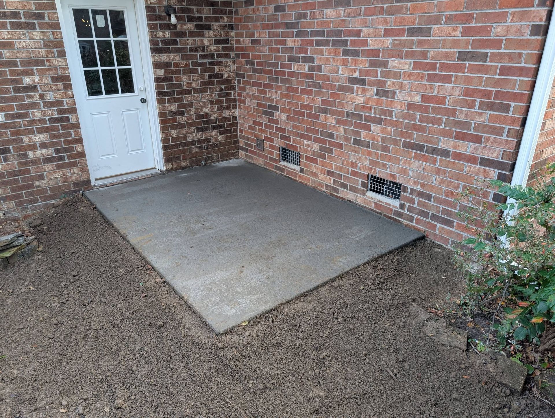 Concrete patio slab beside a white door on a brick wall, surrounded by bare soil and shrubs.