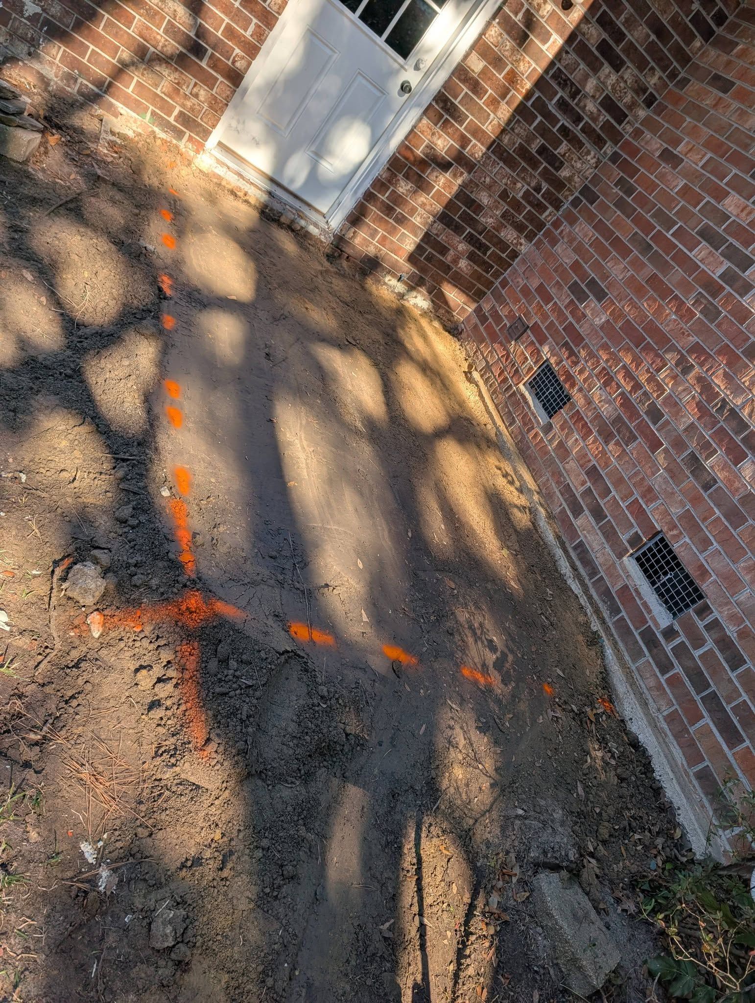 Cracked pavement beside a brick wall, marked with orange spray paint and tree shadows.