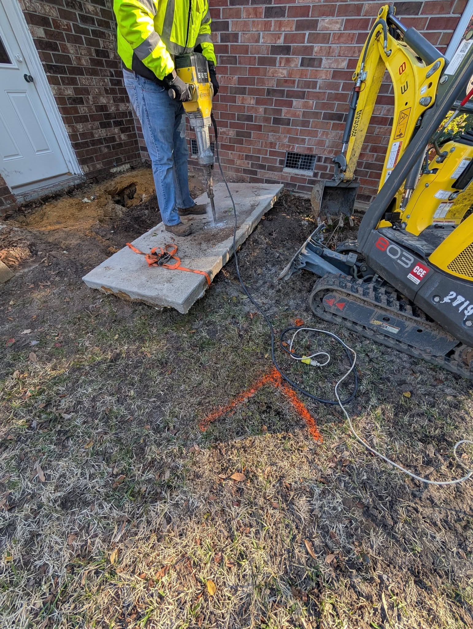Worker using a jackhammer on a concrete slab beside a brick wall and excavator.