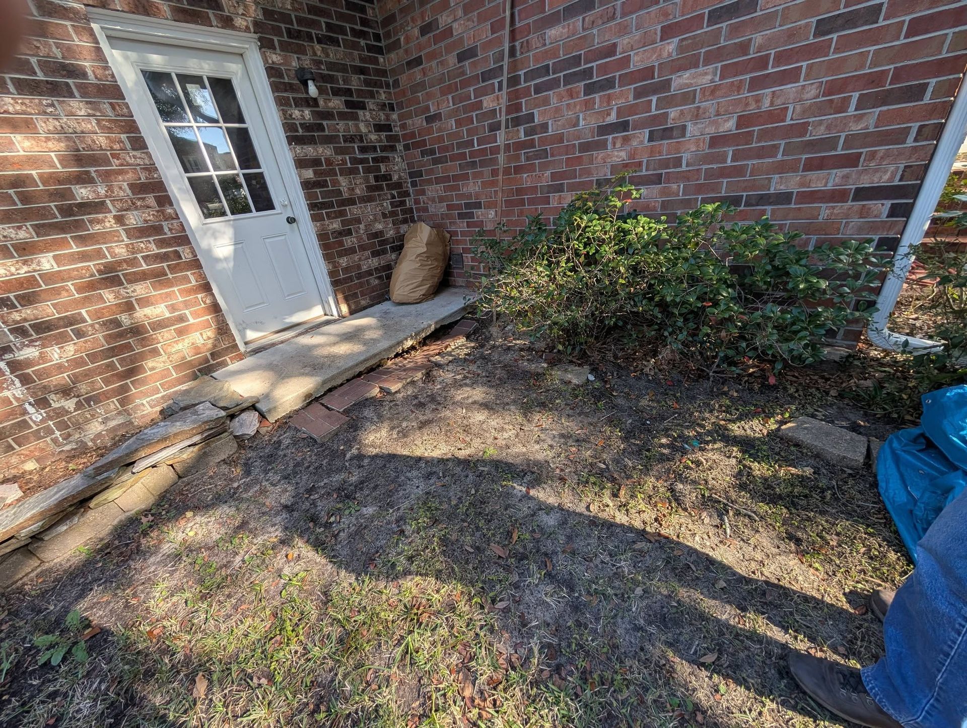Backyard patio beside brick house with white door, shrub, and blue tarp on the right, in bright sunlight