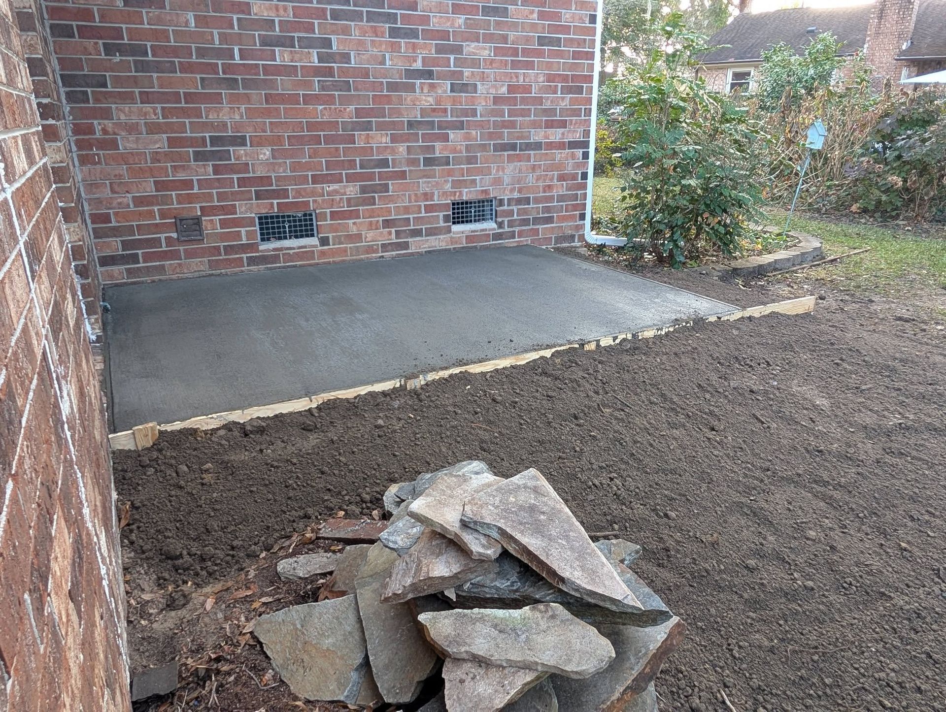 Backyard beside a brick house with a concrete slab and a pile of rocks on bare ground