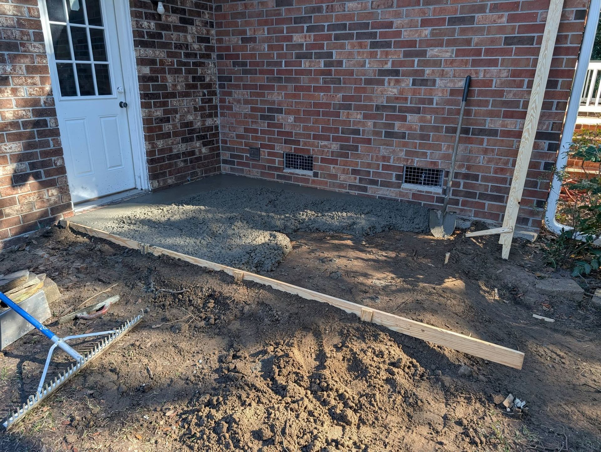 Dirt-covered backyard patio beside a brick house, with a white door and a wooden board on the ground.