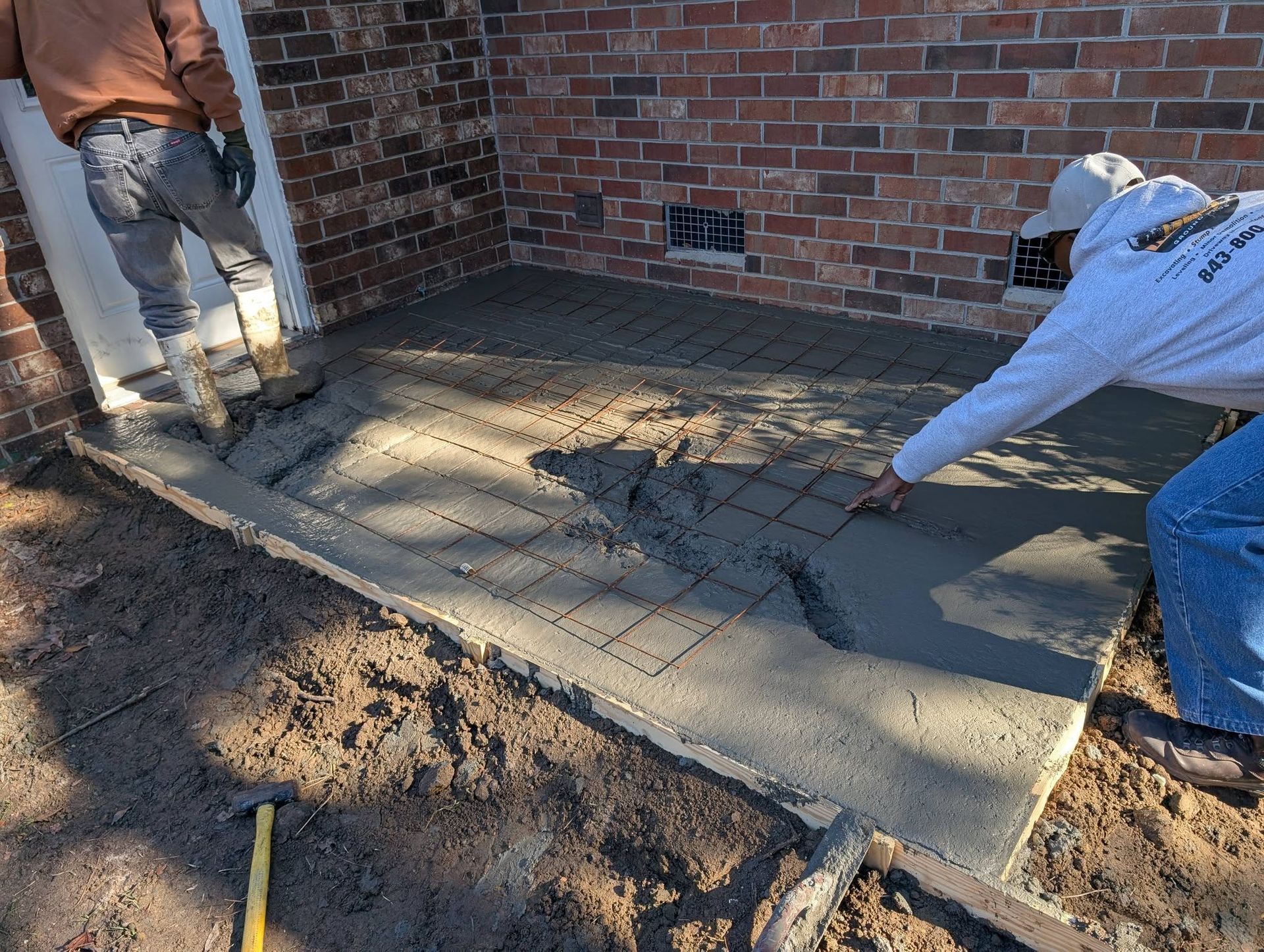 Workers smoothing fresh concrete patio beside a brick wall during construction
