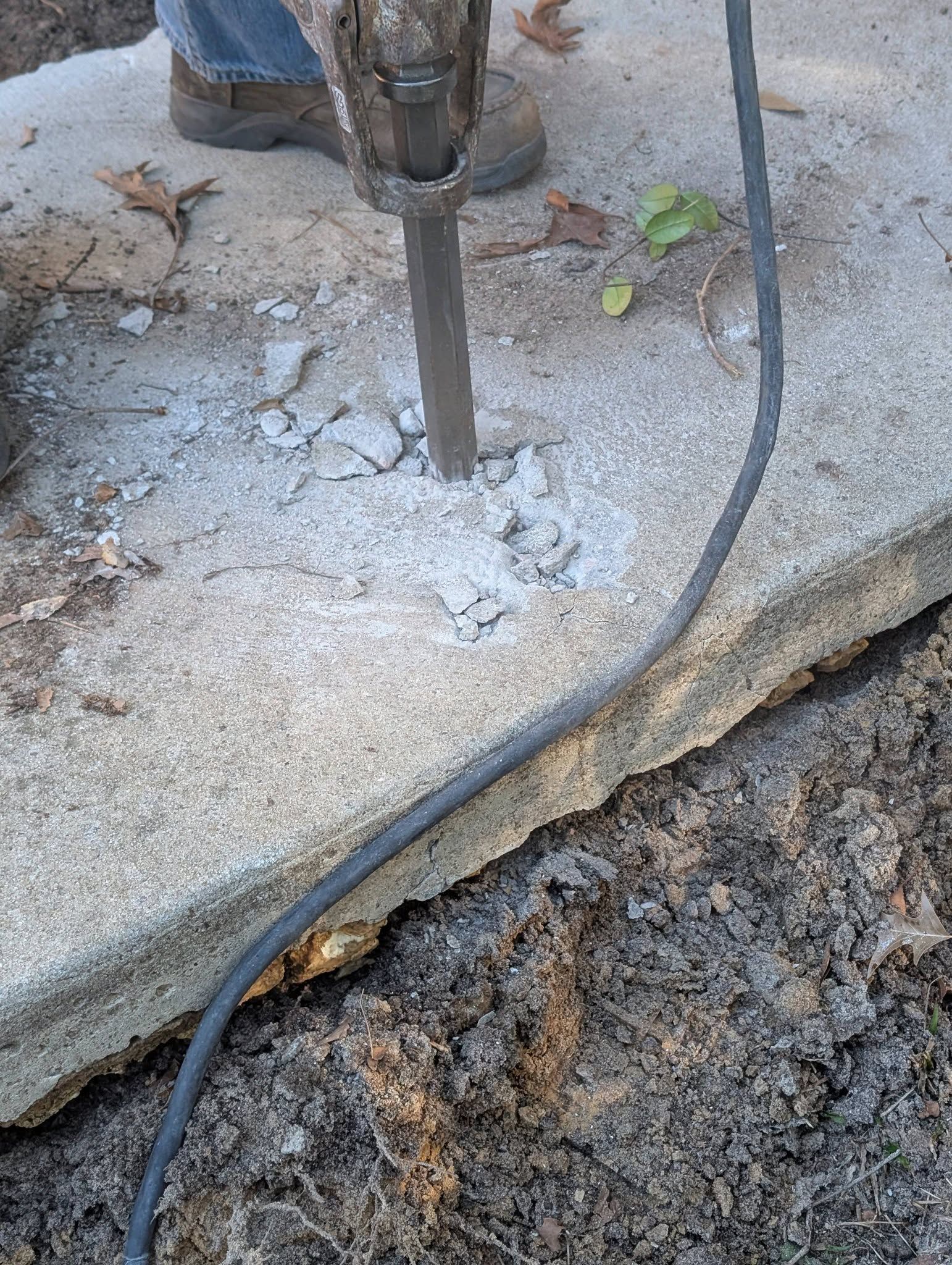 Jackhammer breaking concrete at the edge of a damaged sidewalk, with a cable beside it.