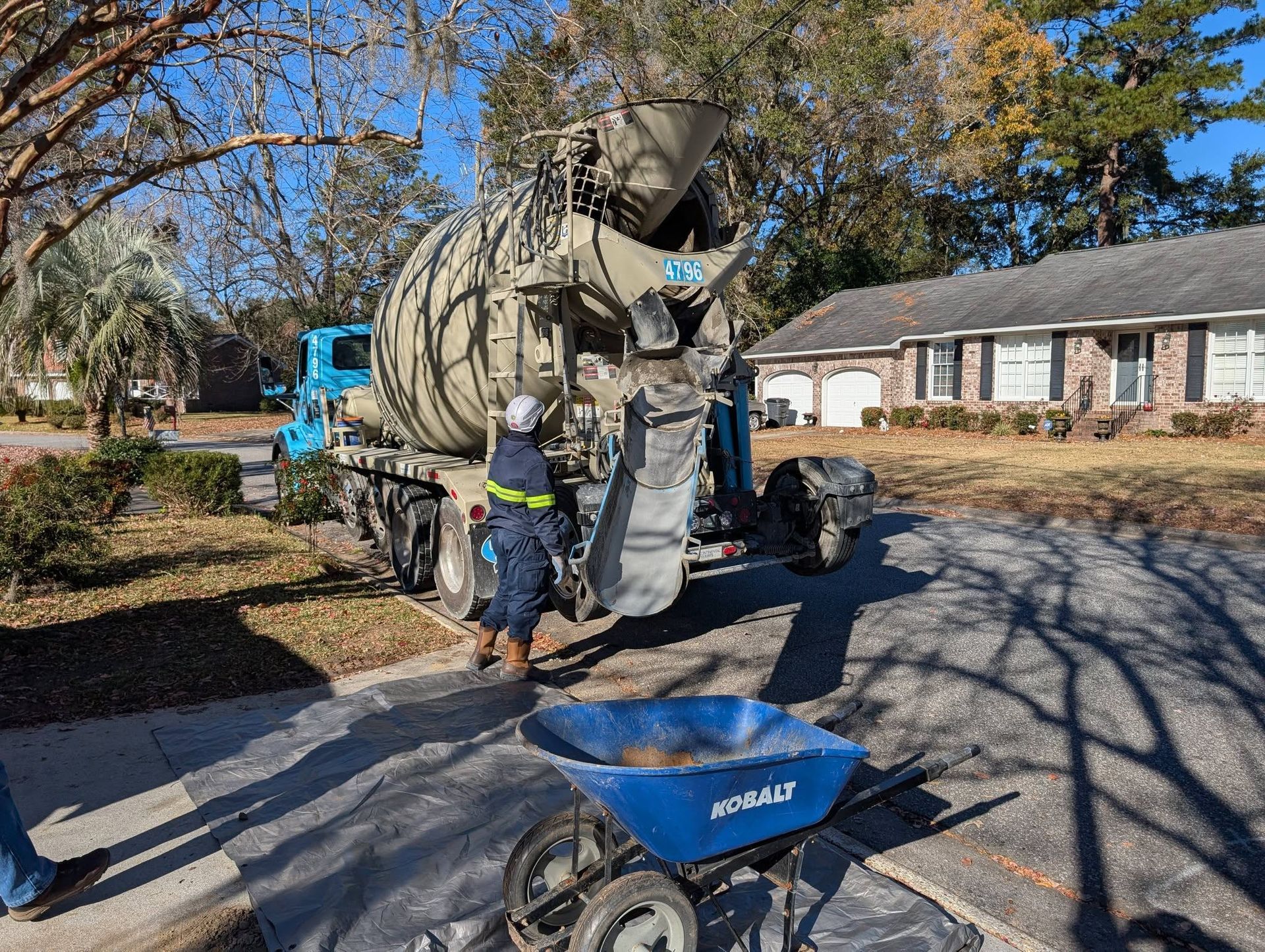 Worker guides a cement mixer truck on a residential street with a blue wheelbarrow in the foreground.