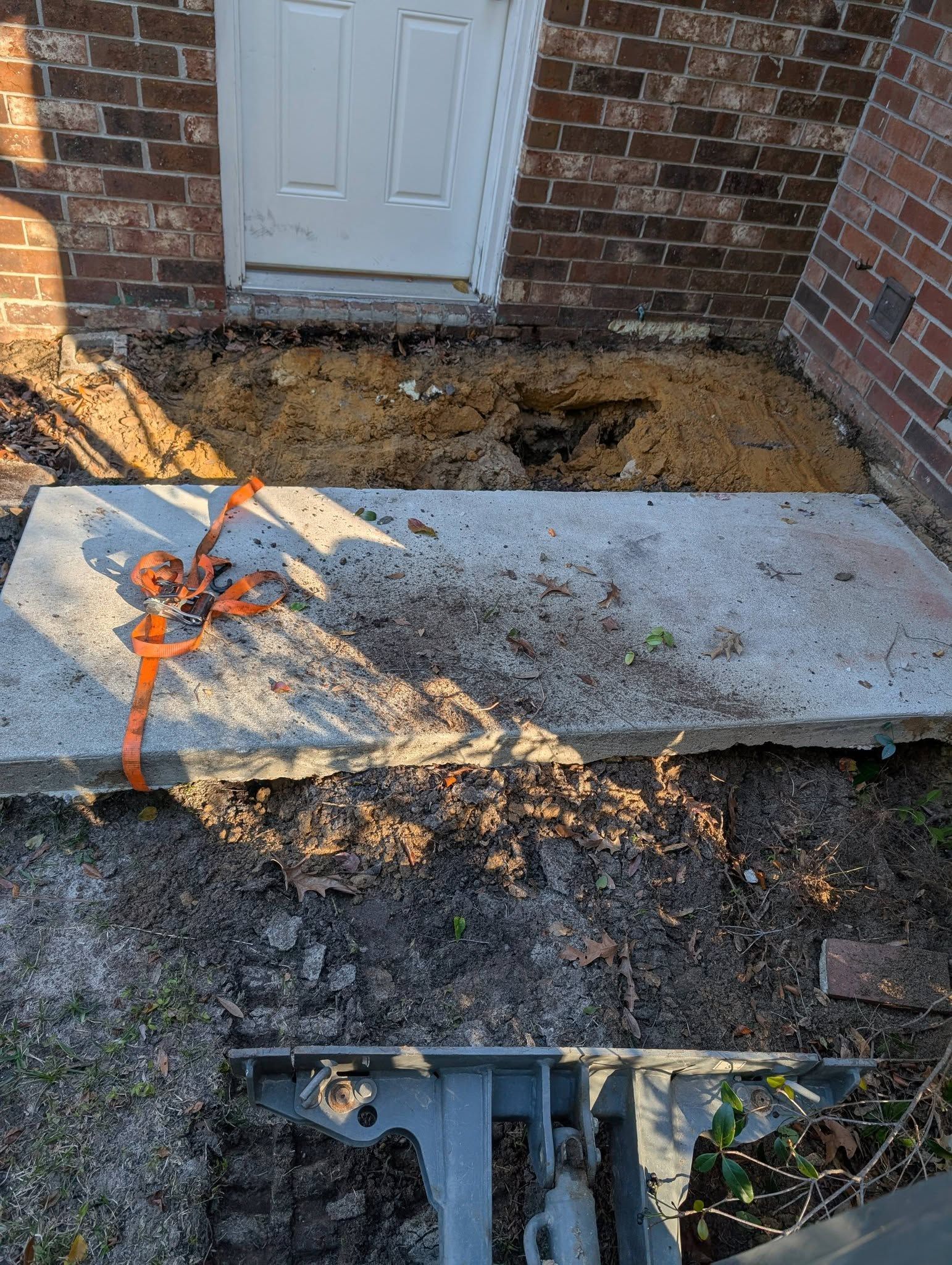 Damaged concrete walkway by a brick wall and white door, with exposed dirt and orange marker paint