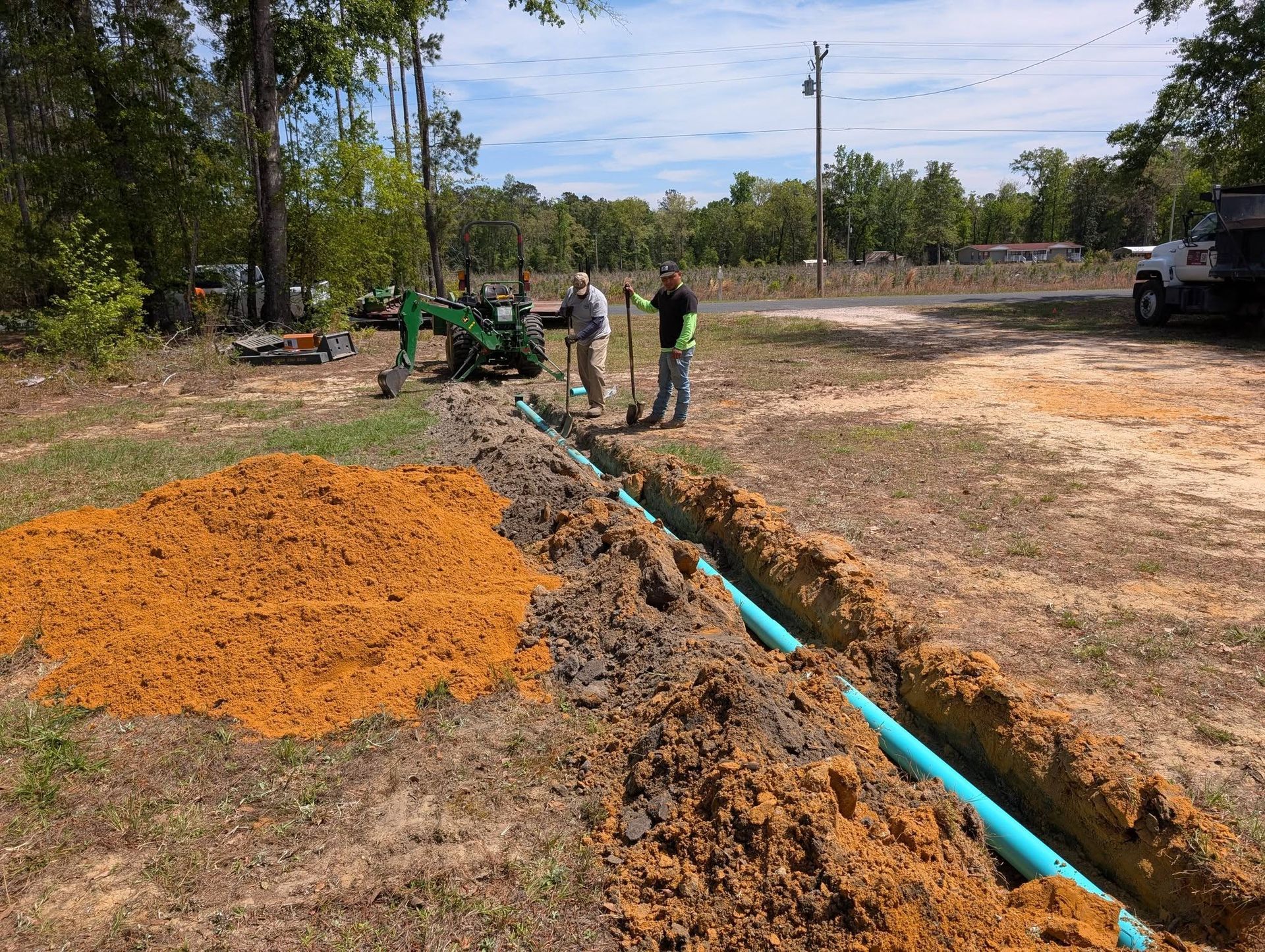 Workers install a blue pipe in a trench beside piles of orange gravel at a roadside construction site.