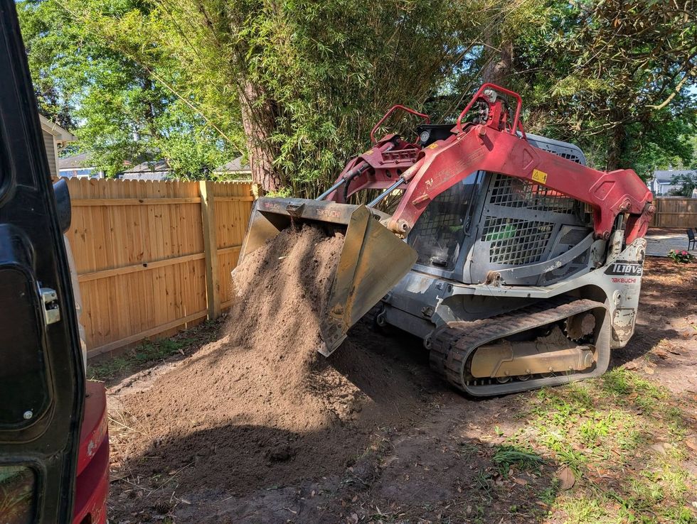 Red skid steer dumping a load of dirt in a backyard beside a wooden fence and trees