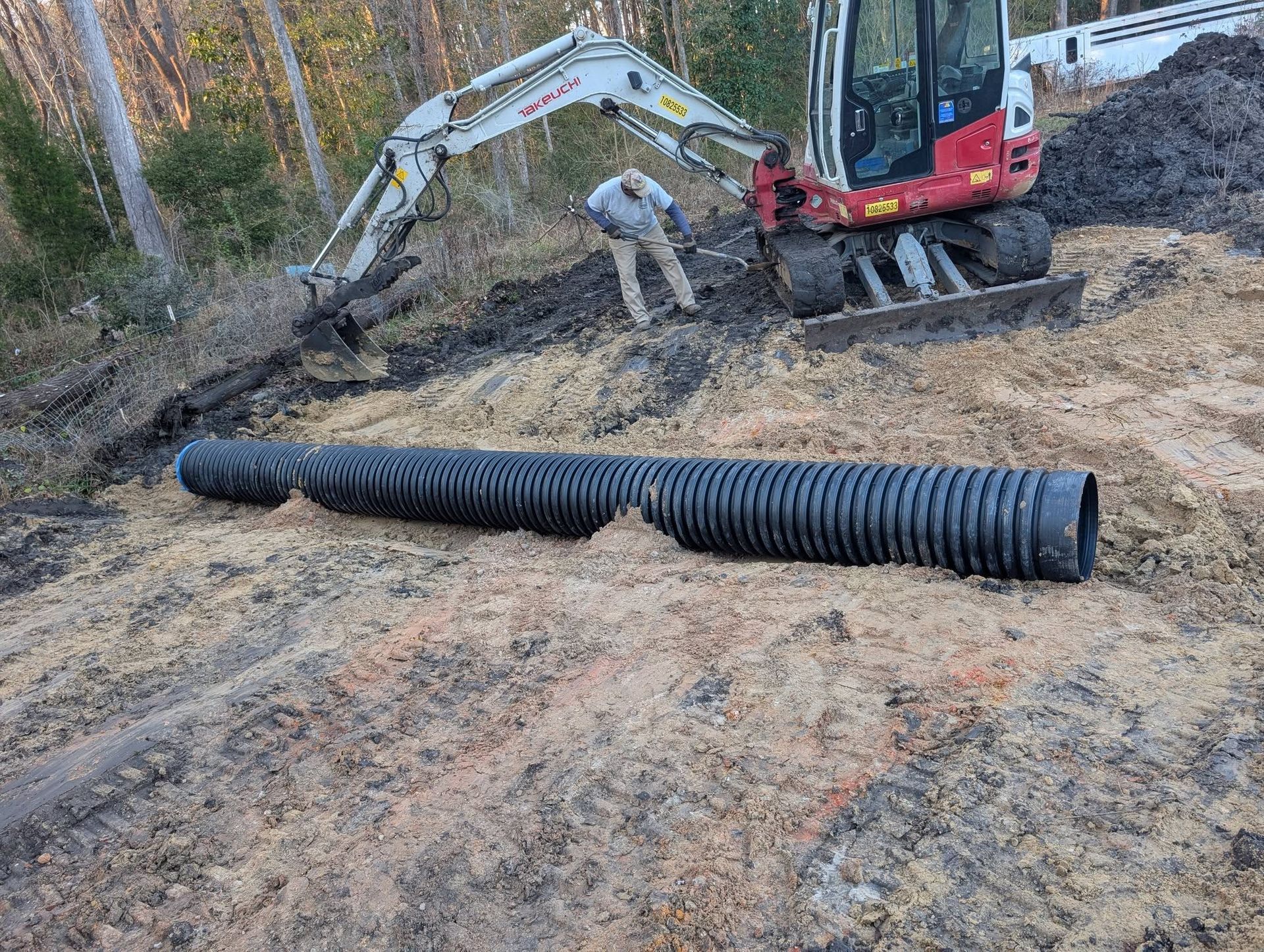 Construction site with workers installing a large black corrugated drainage pipe beside an excavator.