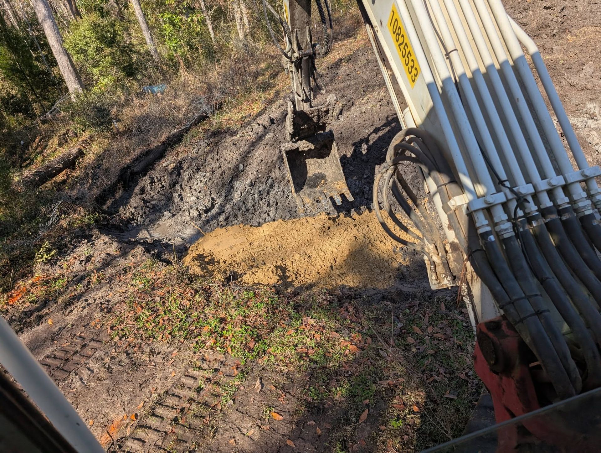 Excavator bucket beside a trench, with black and white pipes stacked on the right in a muddy worksite