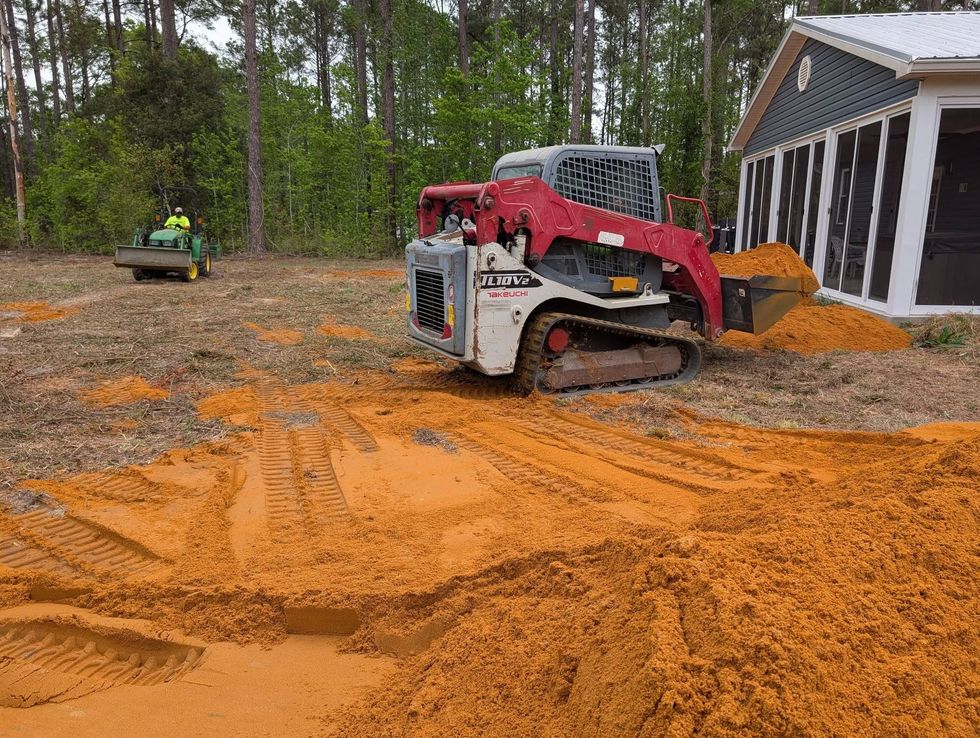 Red and white skid steer spreading orange sand beside a house in a wooded yard.
