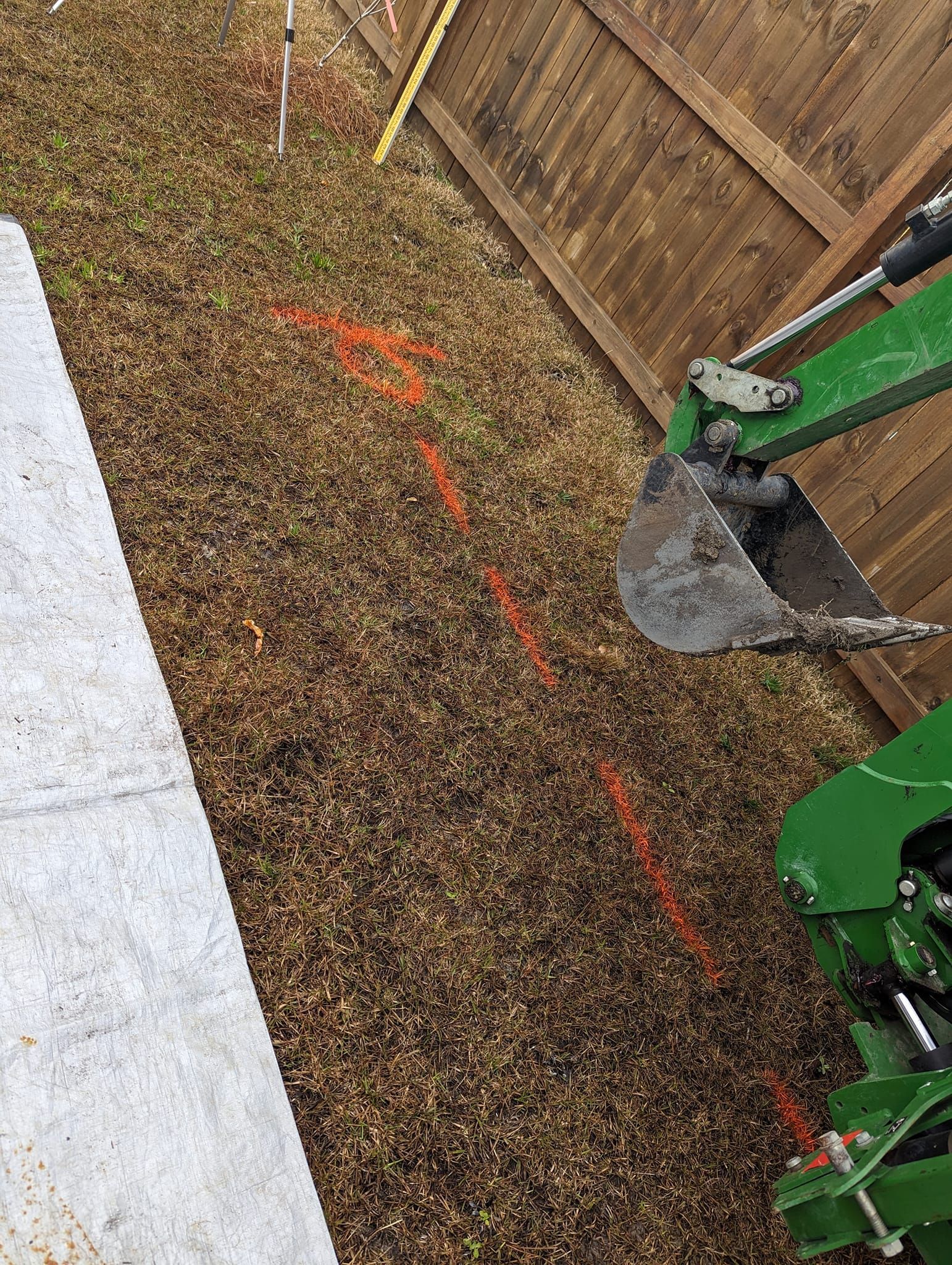 Excavator bucket digging along a gravel edge beside a fence and paving stones
