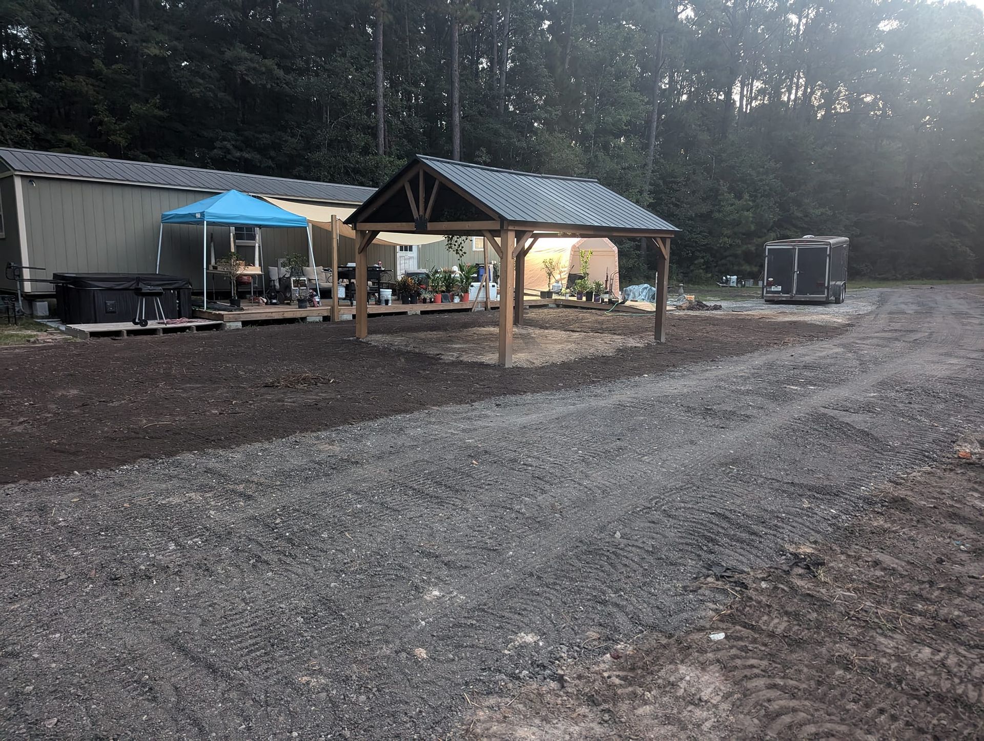 Rustic campsite with a small pavilion, blue canopy, and gravel road in a wooded area at dusk
