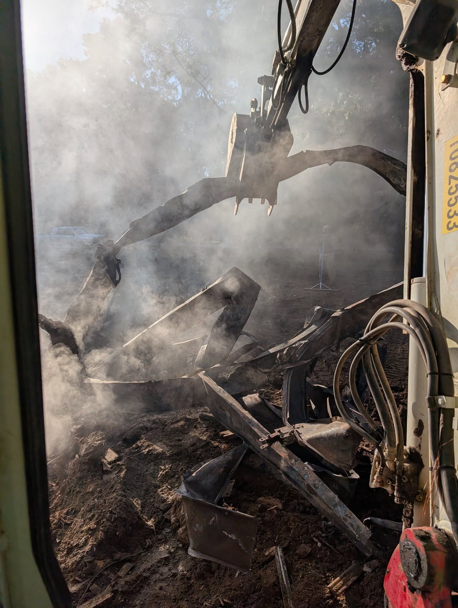 Smoke and flames engulf debris as an excavator works in a fire-ravaged area.