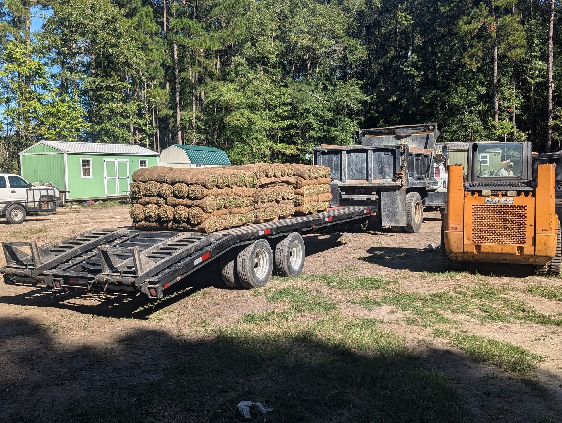 Flatbed trailer loaded with hay bales beside an orange forklift in a wooded yard