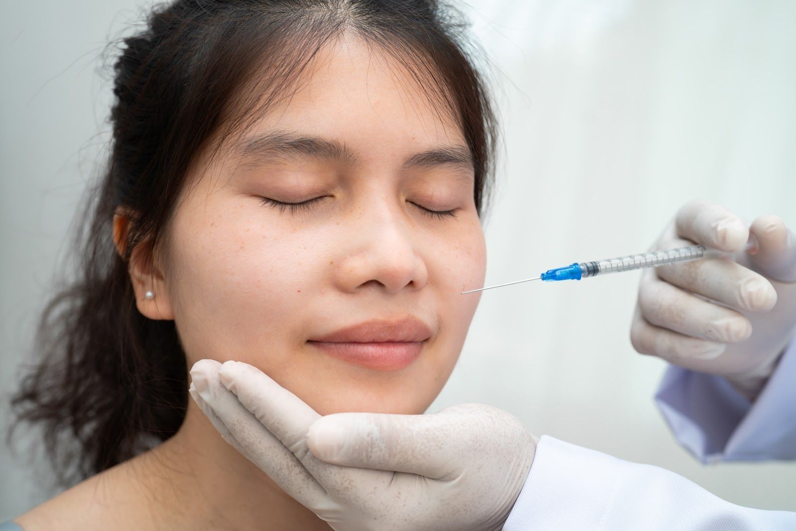 Woman receiving a cosmetic facial injection while holding her chin, eyes closed, in a clinical setting