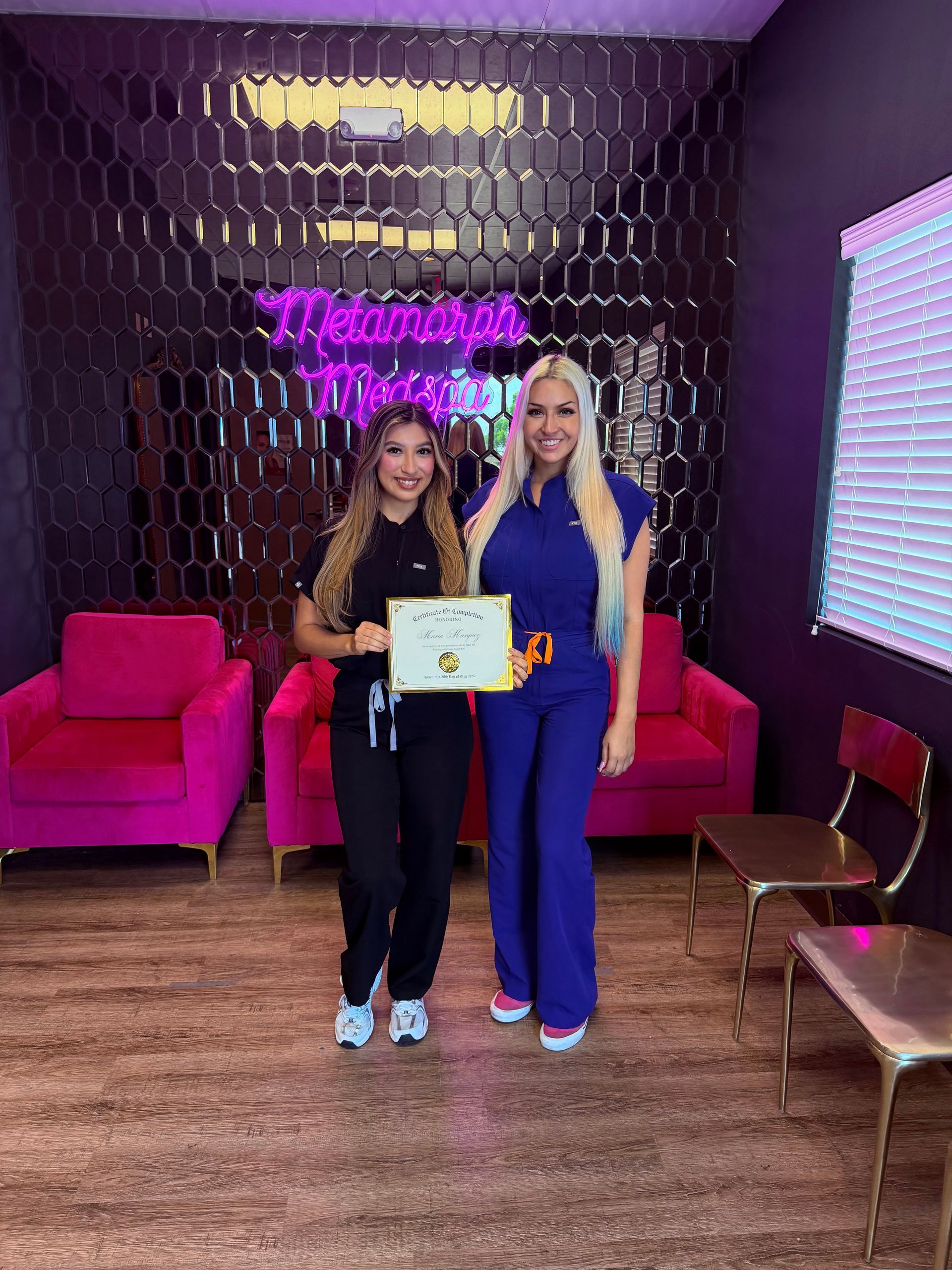 Two women holding a certificate in a purple lounge with pink chairs and decorative wall lighting.