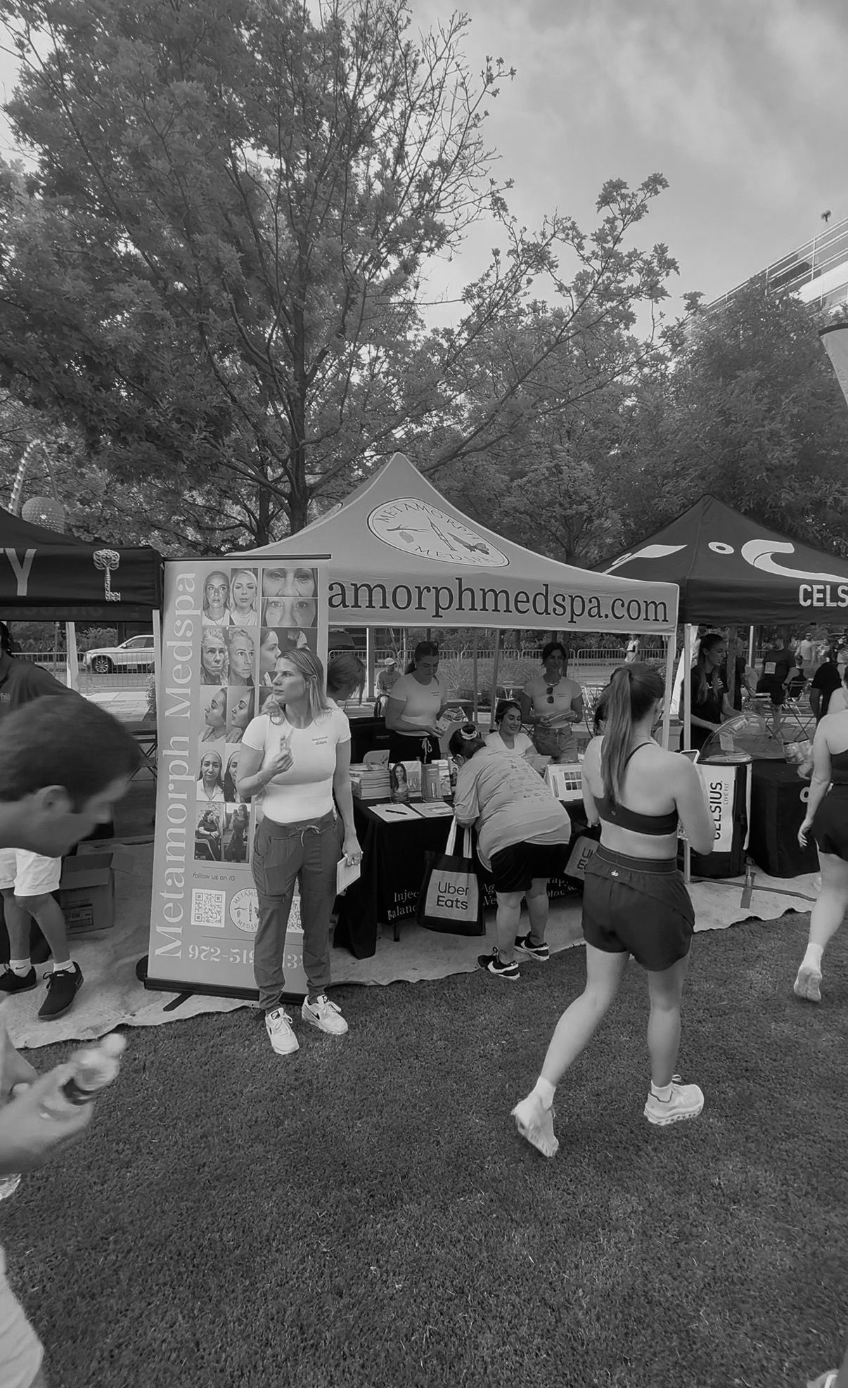 People posing at an outdoor booth with a banner, trees and tents in the background.