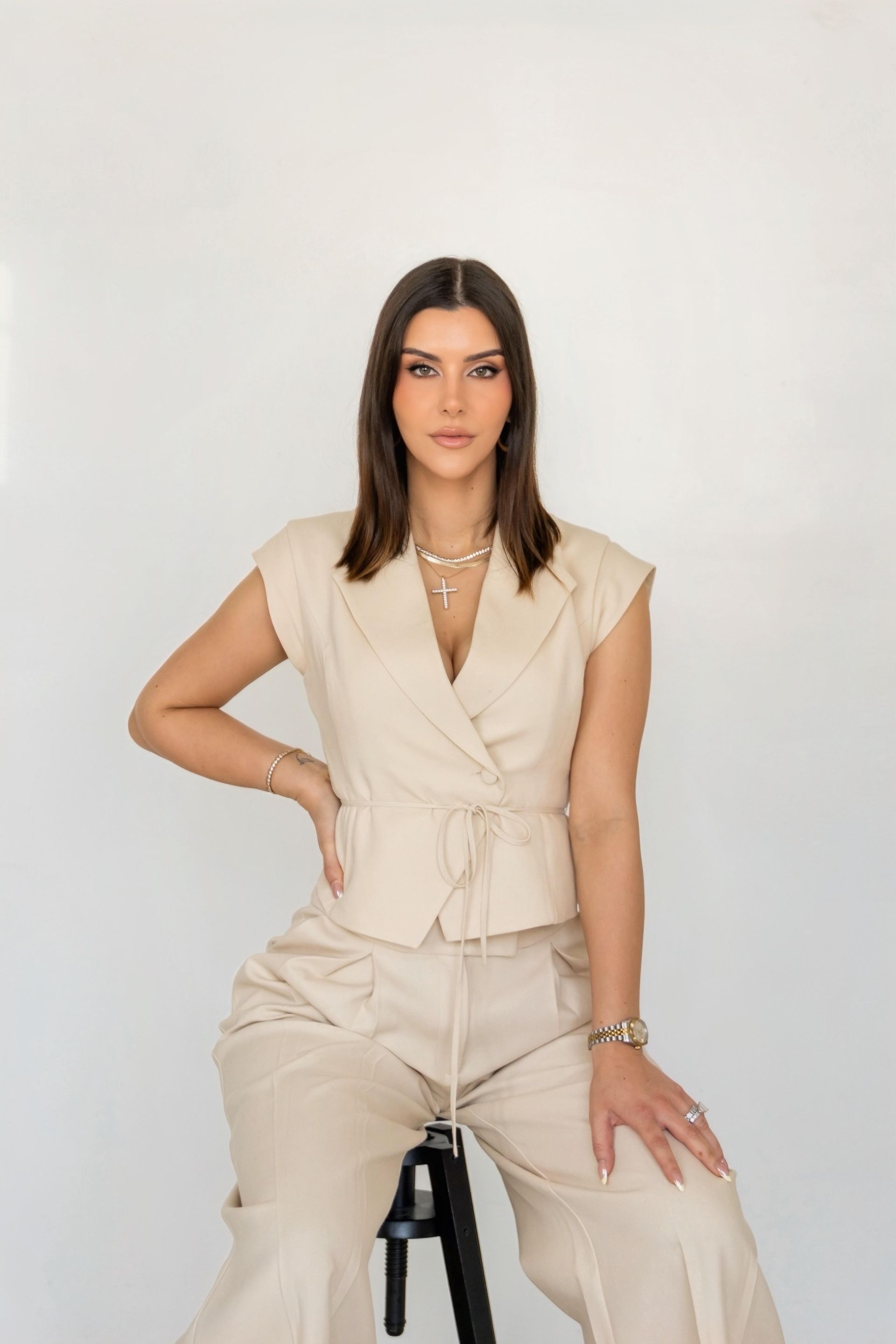 Woman seated on a stool in a cream outfit against a plain white background.