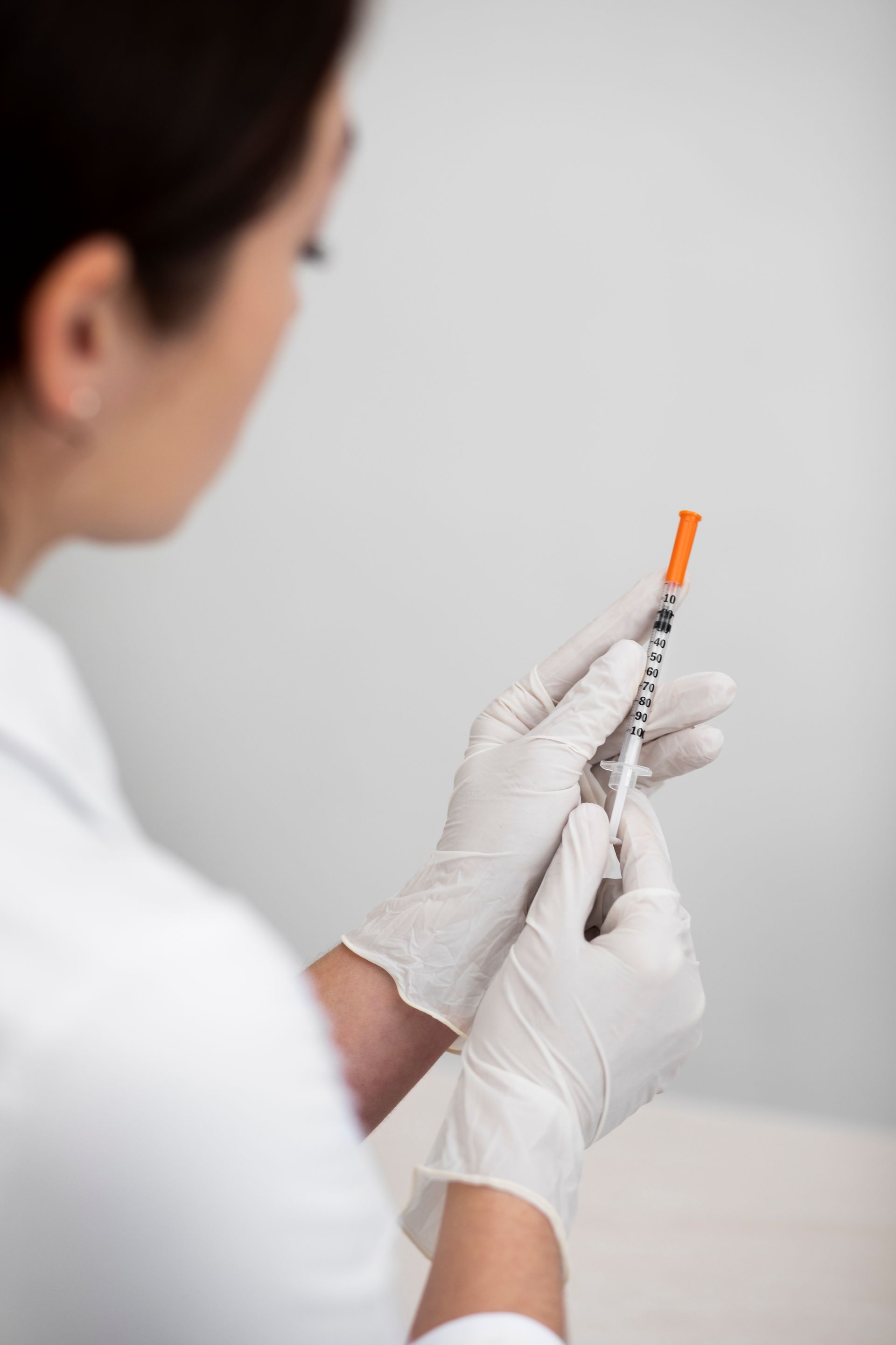 Gloved healthcare worker holding a syringe and orange-capped vial against a plain background