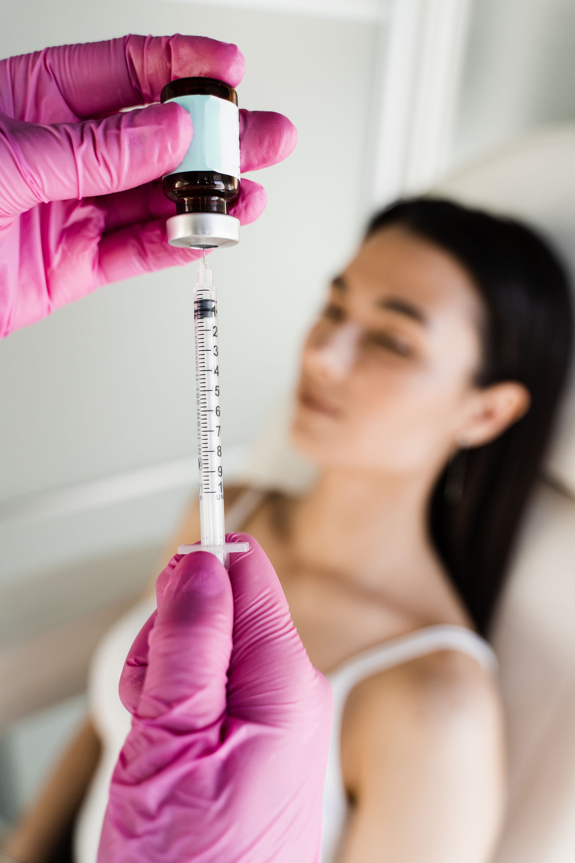 Gloved hands holding a syringe and vial near a blurred patient in a clinical setting