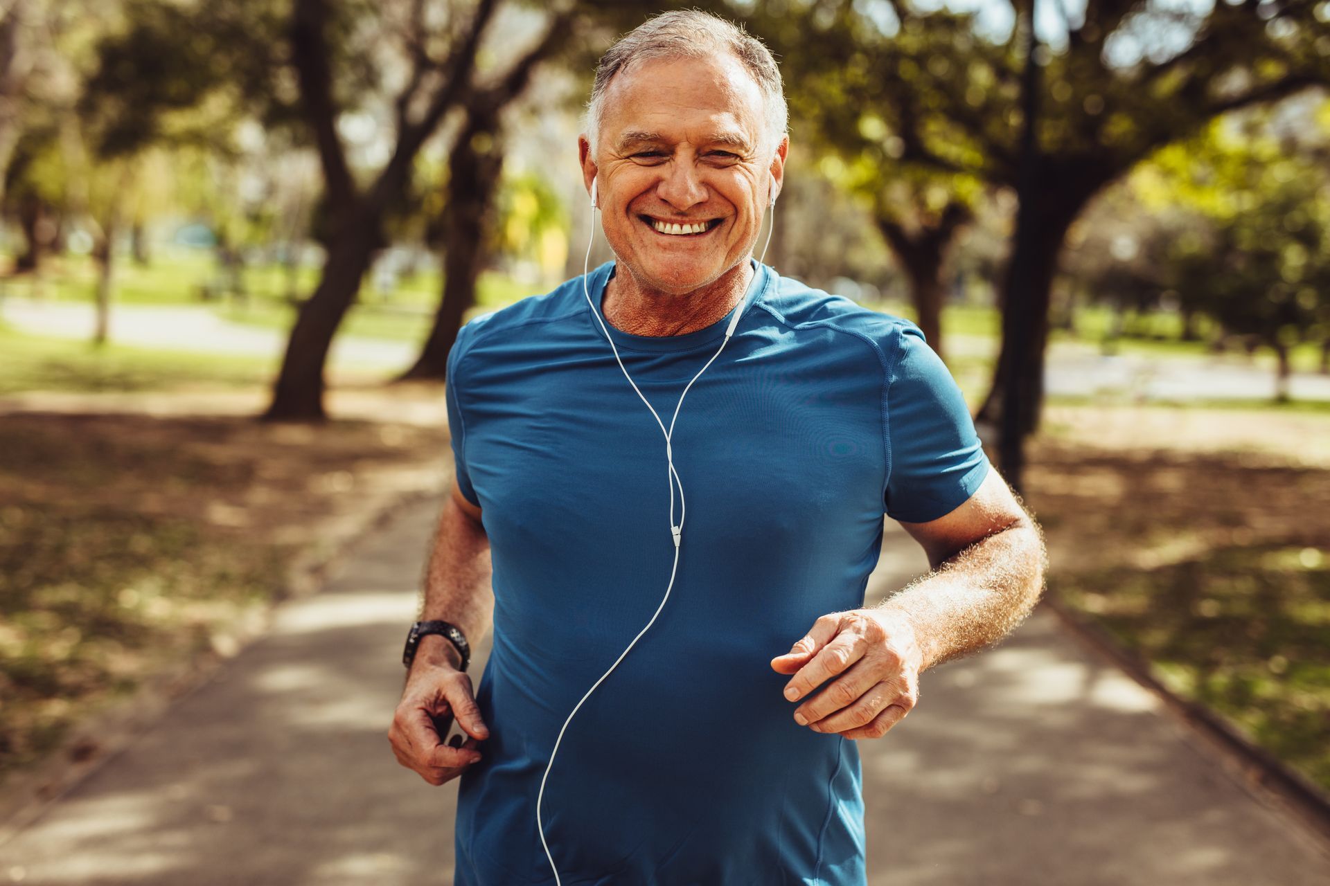Smiling man jogging on a tree-lined path, wearing a blue shirt and earbuds