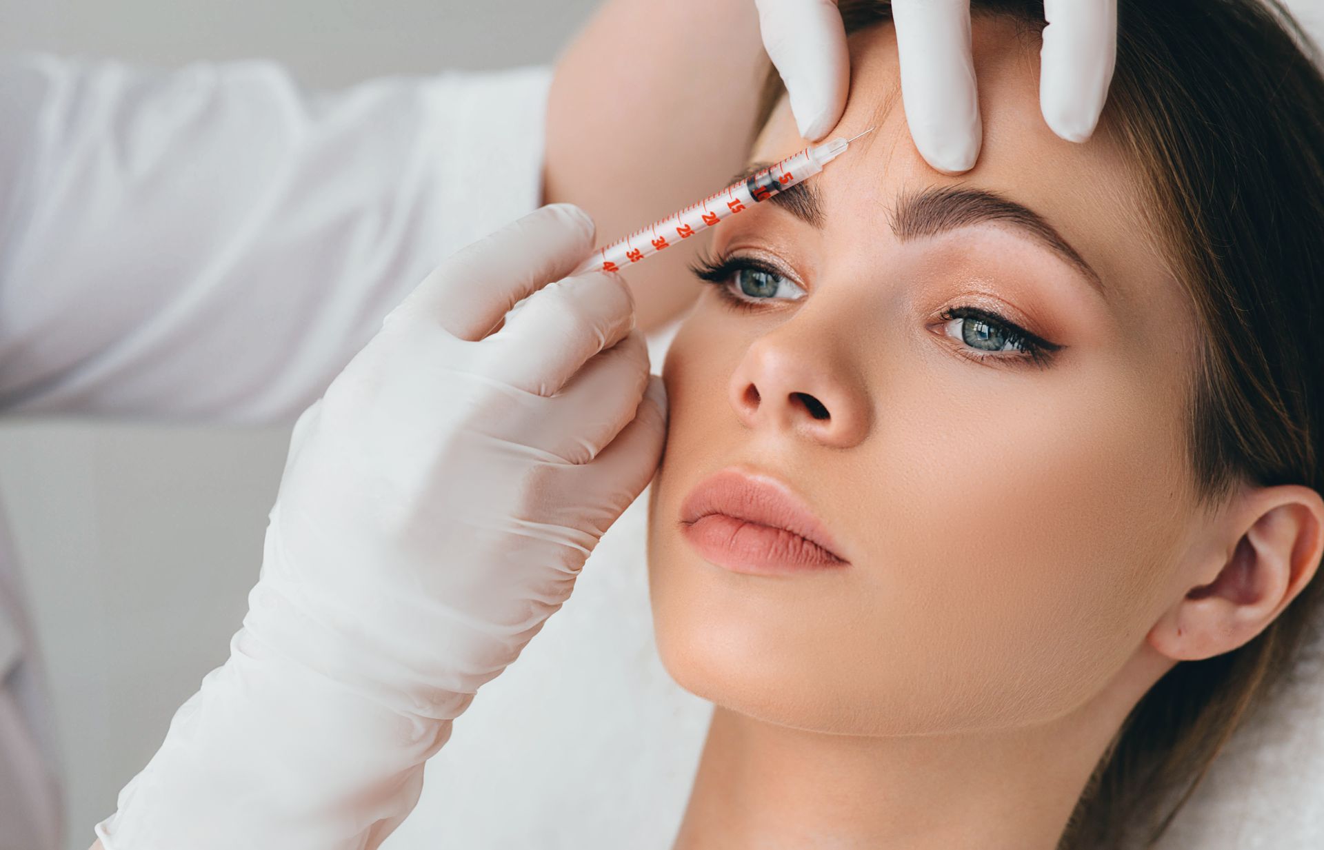Gloved hands inject filler near a woman’s eyebrow as she lies back during a cosmetic procedure
