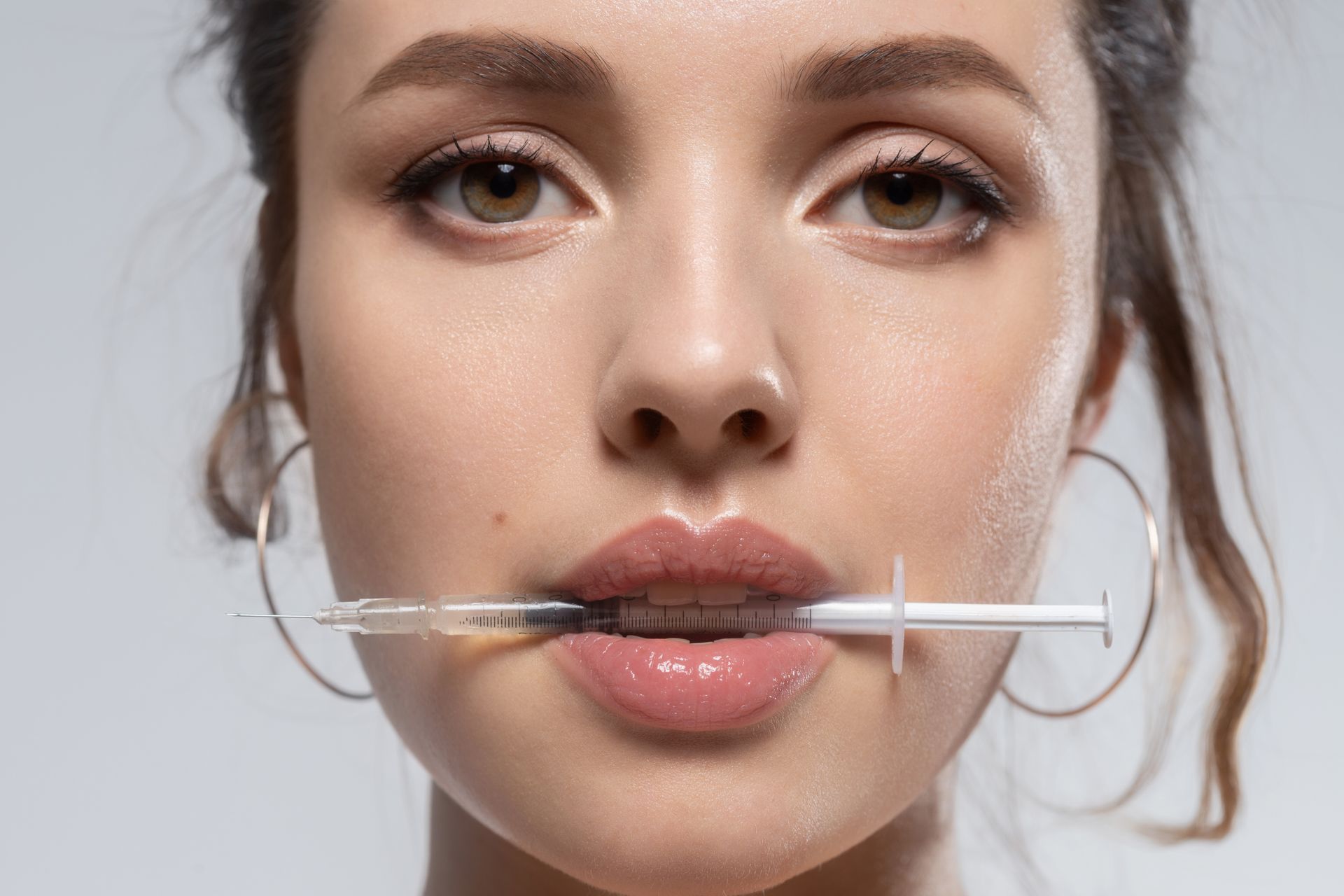 Close-up of a woman holding a syringe near her lips, with hoop earrings and a neutral background