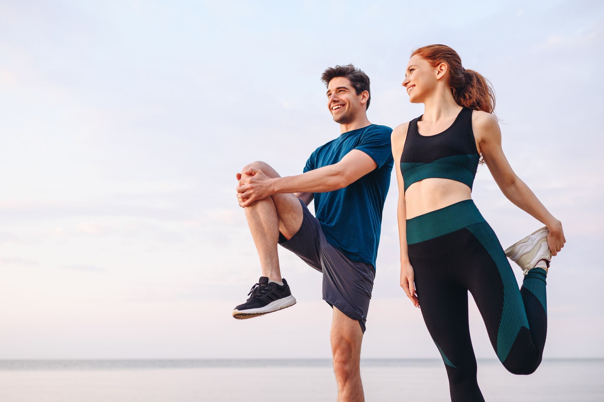 Two people stretching outdoors on a beach at sunset, one in blue and one in black-and-teal workout clothes