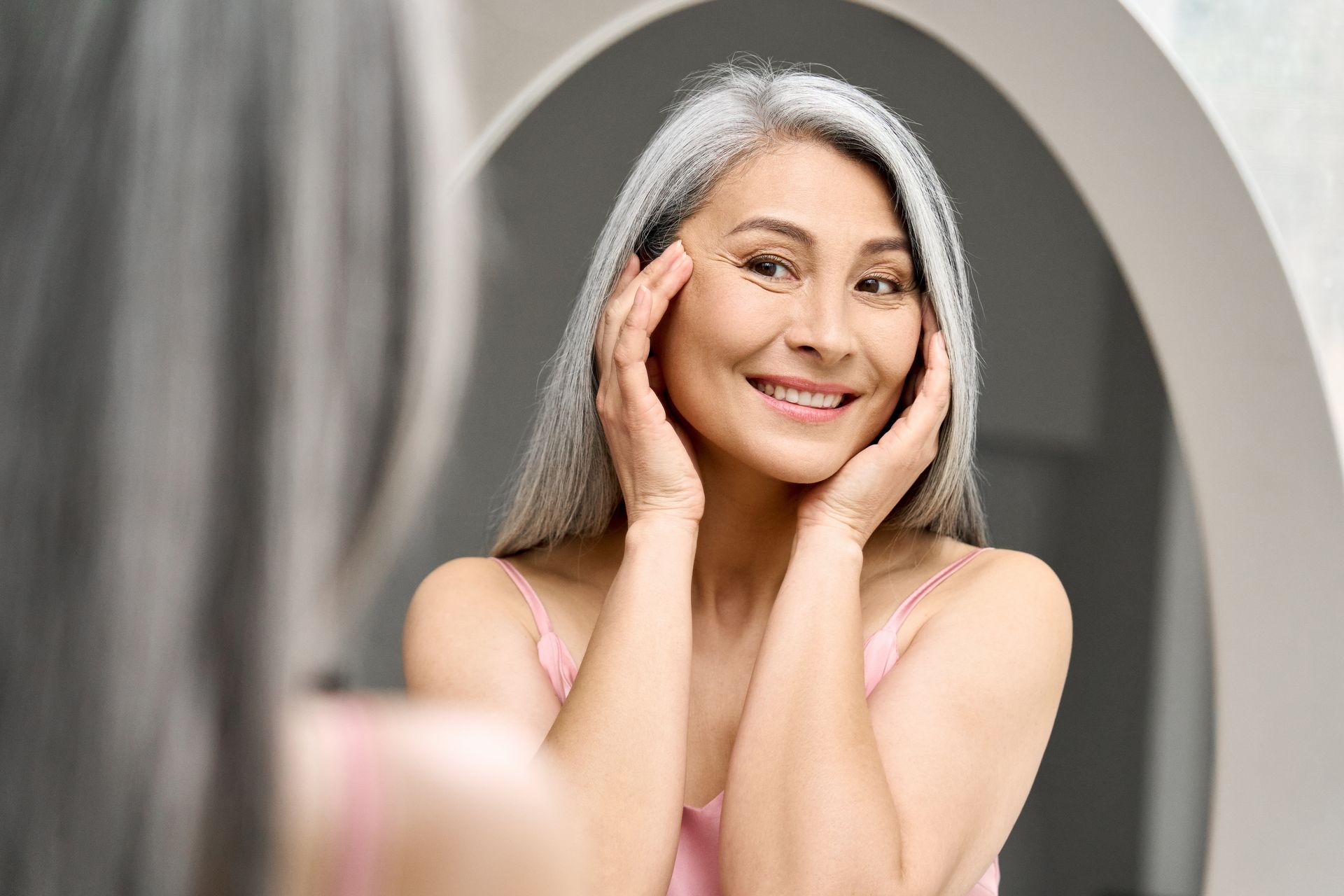 Smiling woman with long gray hair touching her face in front of a mirror