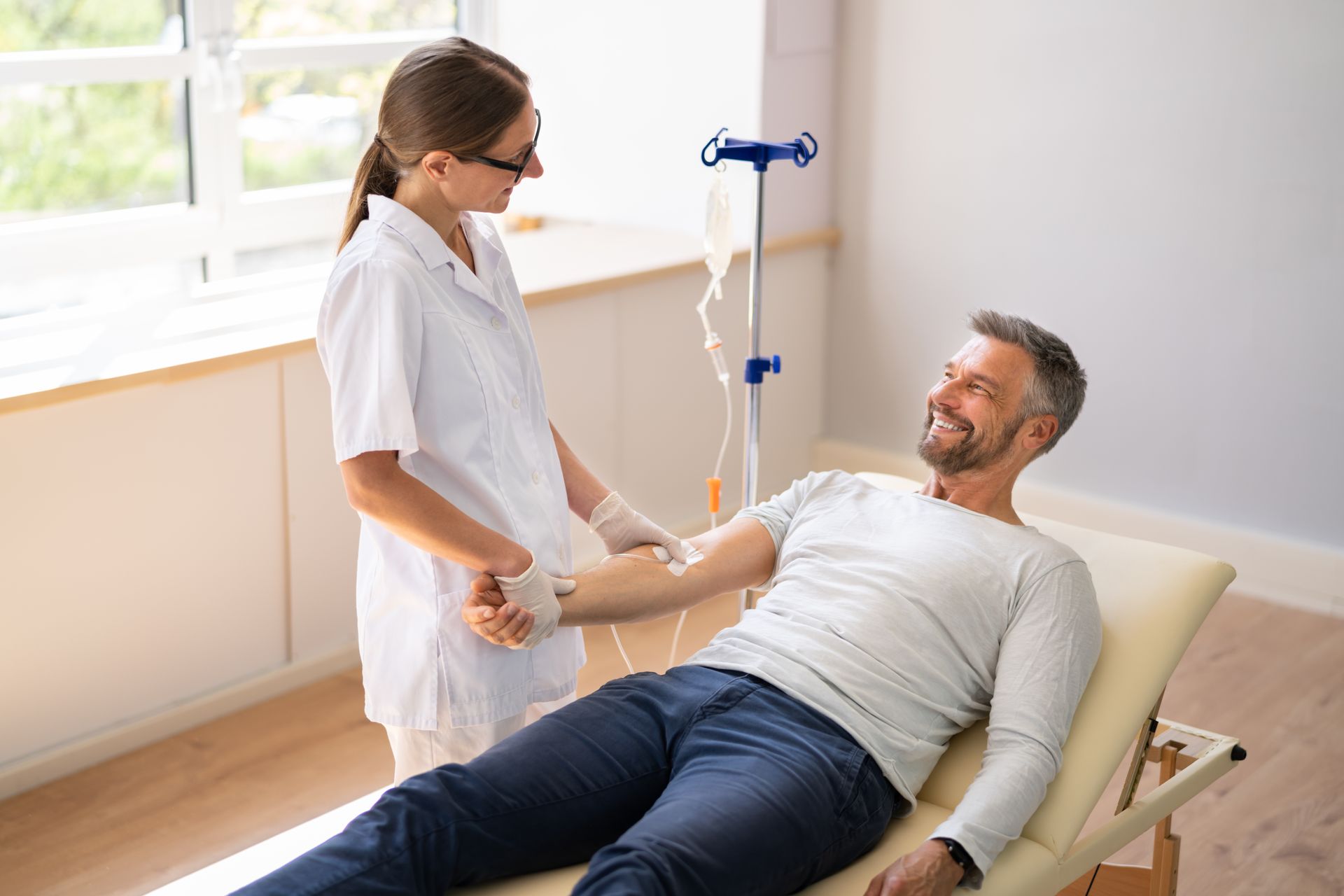 Nurse checking a man’s arm while he lies on a clinic bed beside an IV stand