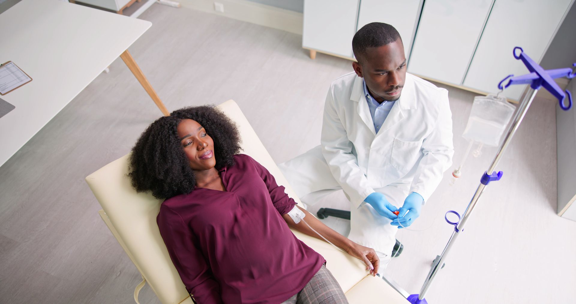 Patient on exam table with doctor in white coat preparing an IV line in a bright clinic room