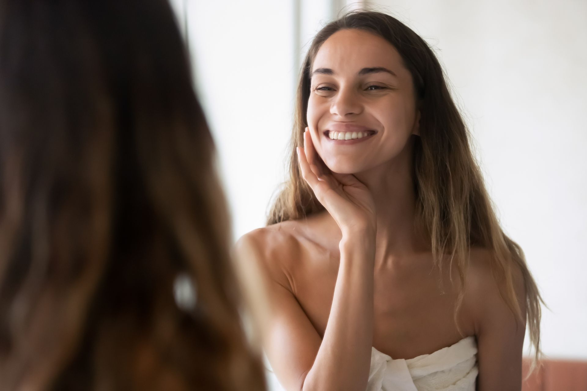 Smiling woman touching her cheek while looking in a mirror, wearing a strapless top.